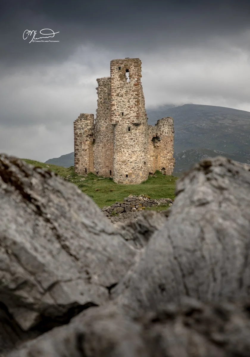Scotland, north coast 500, NC500, Assynt. Ardvrek Castle. An ancient stone castle with a tall central tower, surrounded by grassy land and rocks, under a cloudy sky with mountains in the background.