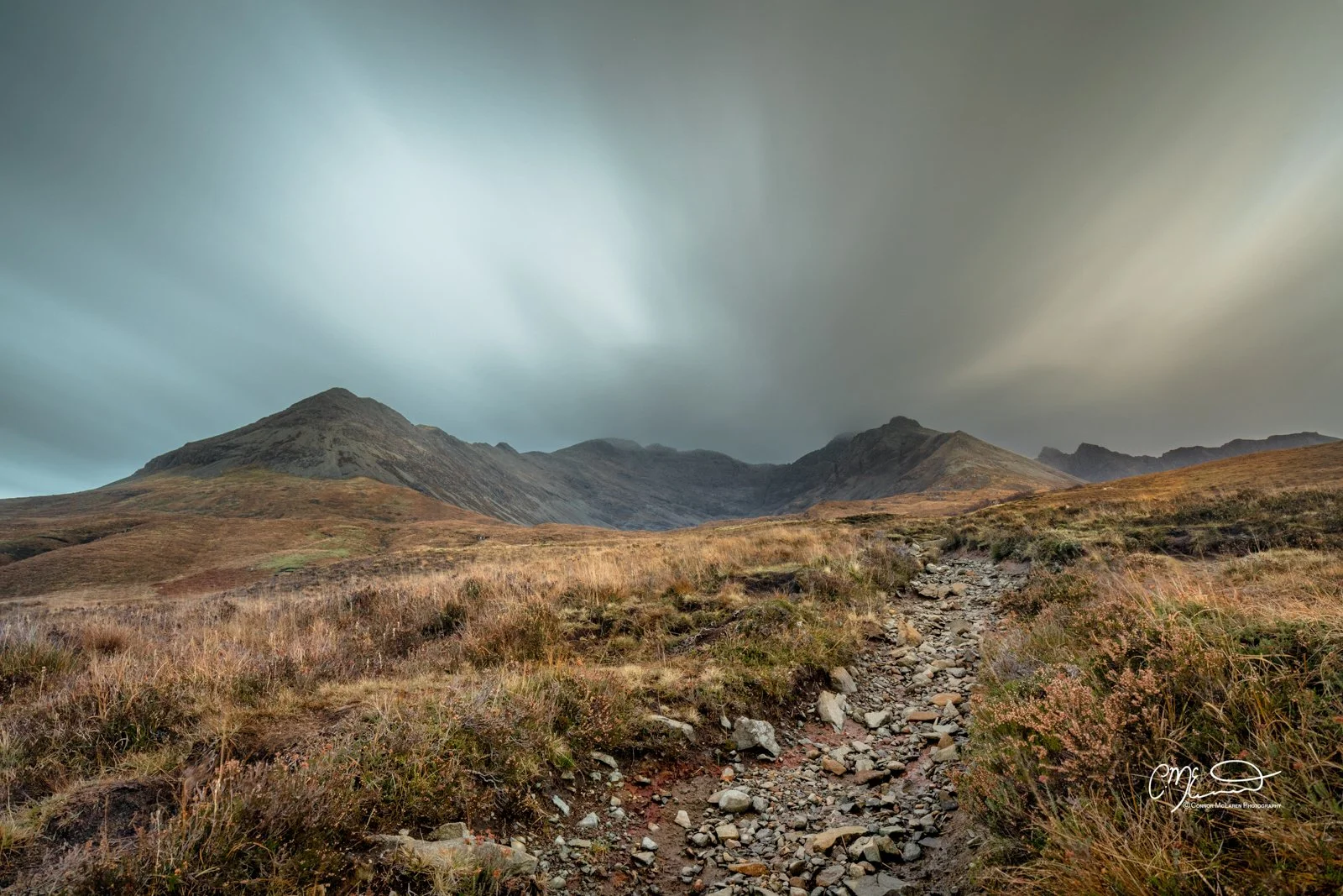 Scotland, Isle of Skye, Fairy Pools. A rugged mountain landscape with a rocky trail leading toward distant peaks under a cloudy sky.