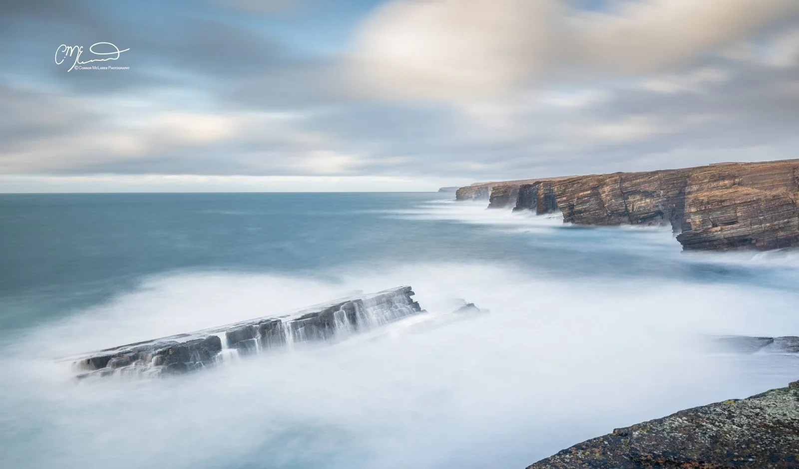 Yesnaby, rainbow, Orkney, Scotland. Long exposure photograph of rugged cliffs along the ocean coast during cloudy weather, with waves crashing against rocks and no visible people.