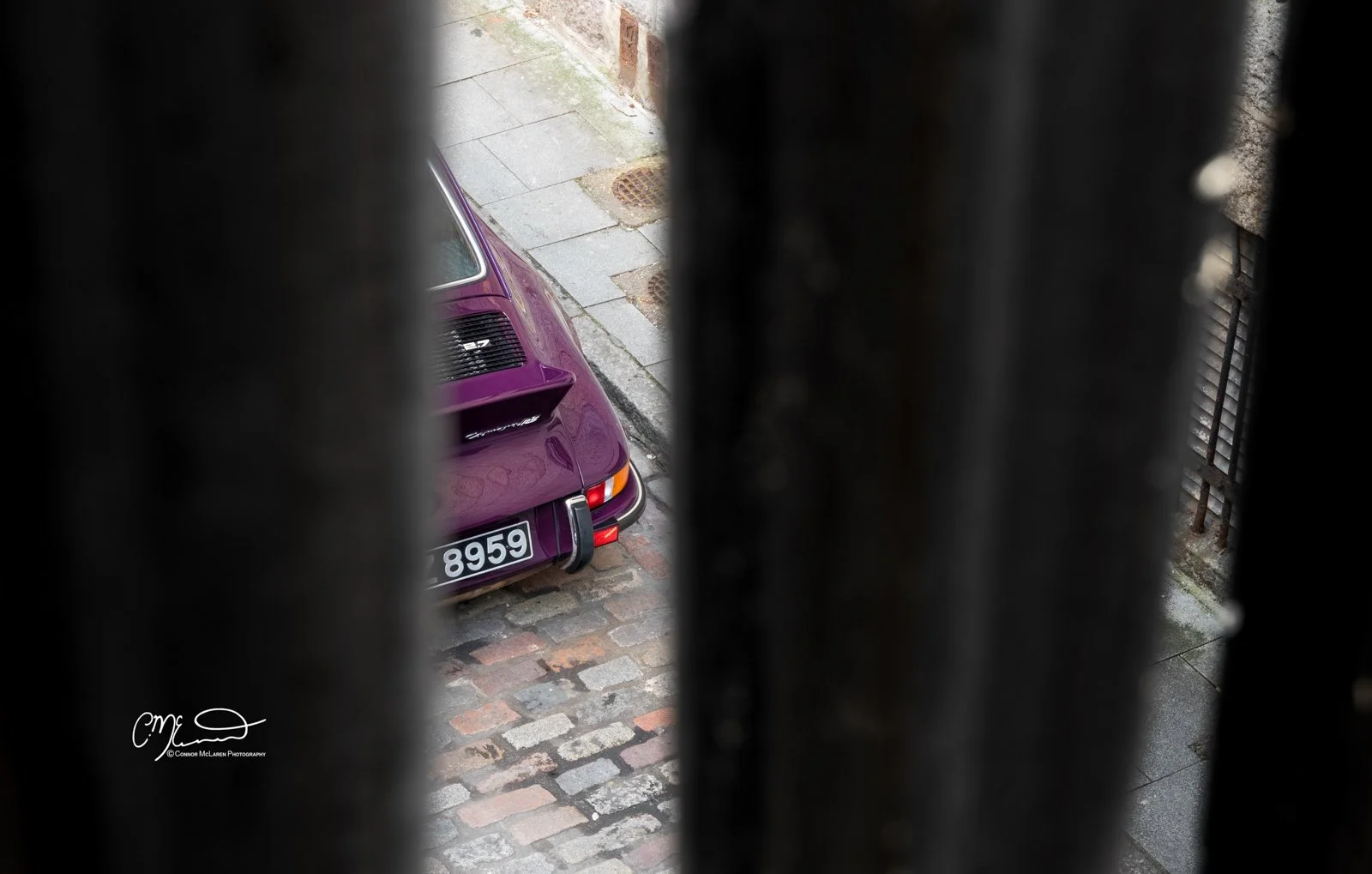 Porsche 911, 2.7RS. View of a purple vintage Porsche car with the license plate 8959, seen through black metal bars from a window, with cobblestone pavement and a sidewalk in the background.