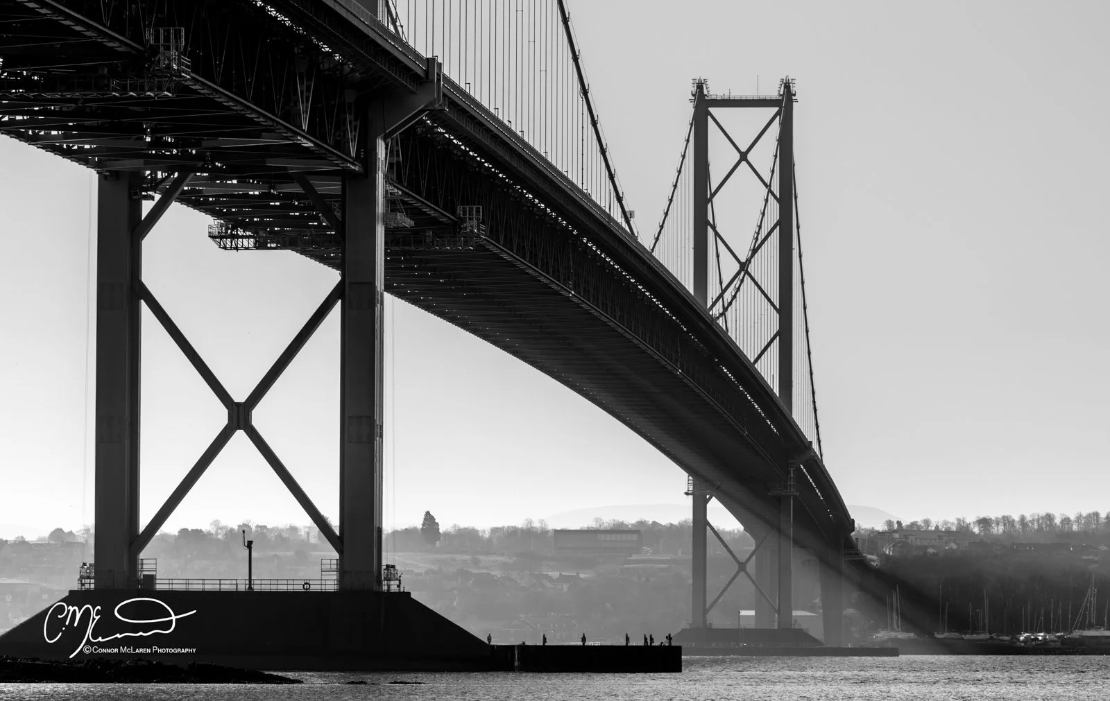 Scotland, Edinburgh Forth Bridge. Black and white photo of a large suspension bridge extending over water, with a distant shoreline and hills in the background.