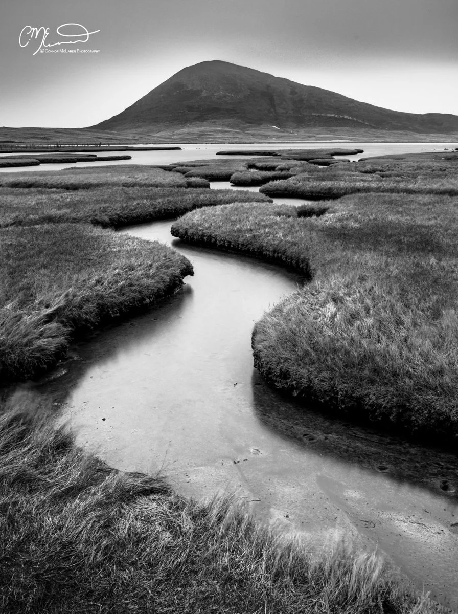 Scotland, Isle of Harris. Black-and-white photograph of a winding river flowing through grassy marshland with a large hill or mountain in the background.