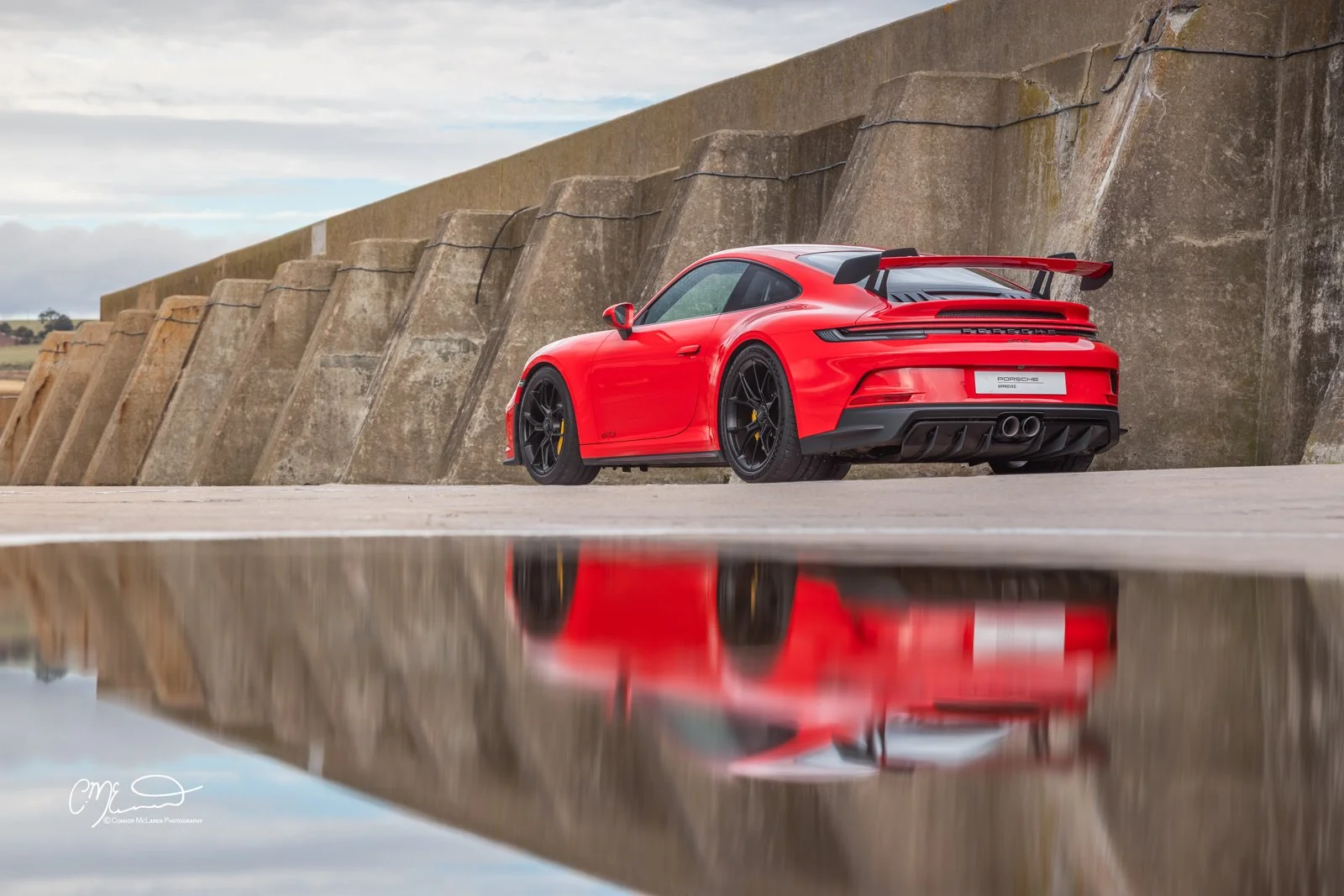 A red Porsche 911 GT3, 992 sports car with black wheels and a rear spoiler parked on a concrete surface, reflected in a puddle, with a large concrete wall and cloudy sky in the background.