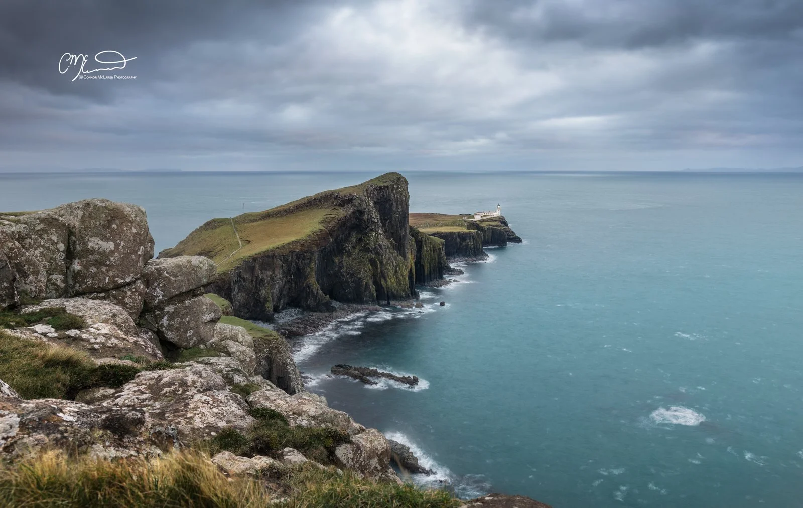 Scotland, Isle of Skye, neist point. Cliffs along a coastline with a lighthouse in the distance under a cloudy sky.