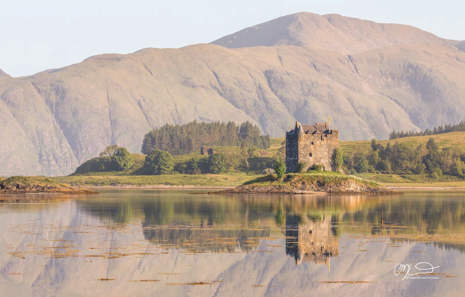 Castle Stalker, Western Highlands, Scotland. A historic castle situated on a small island surrounded by calm water, with lush green trees and hills in the background and mountains in the distance, during daylight.