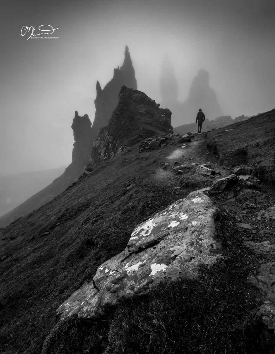Scotland, Isle of Skye old man of storr. A solitary person walking along a rugged, rocky mountain trail with jagged peaks in distance, shrouded in fog in black and white.