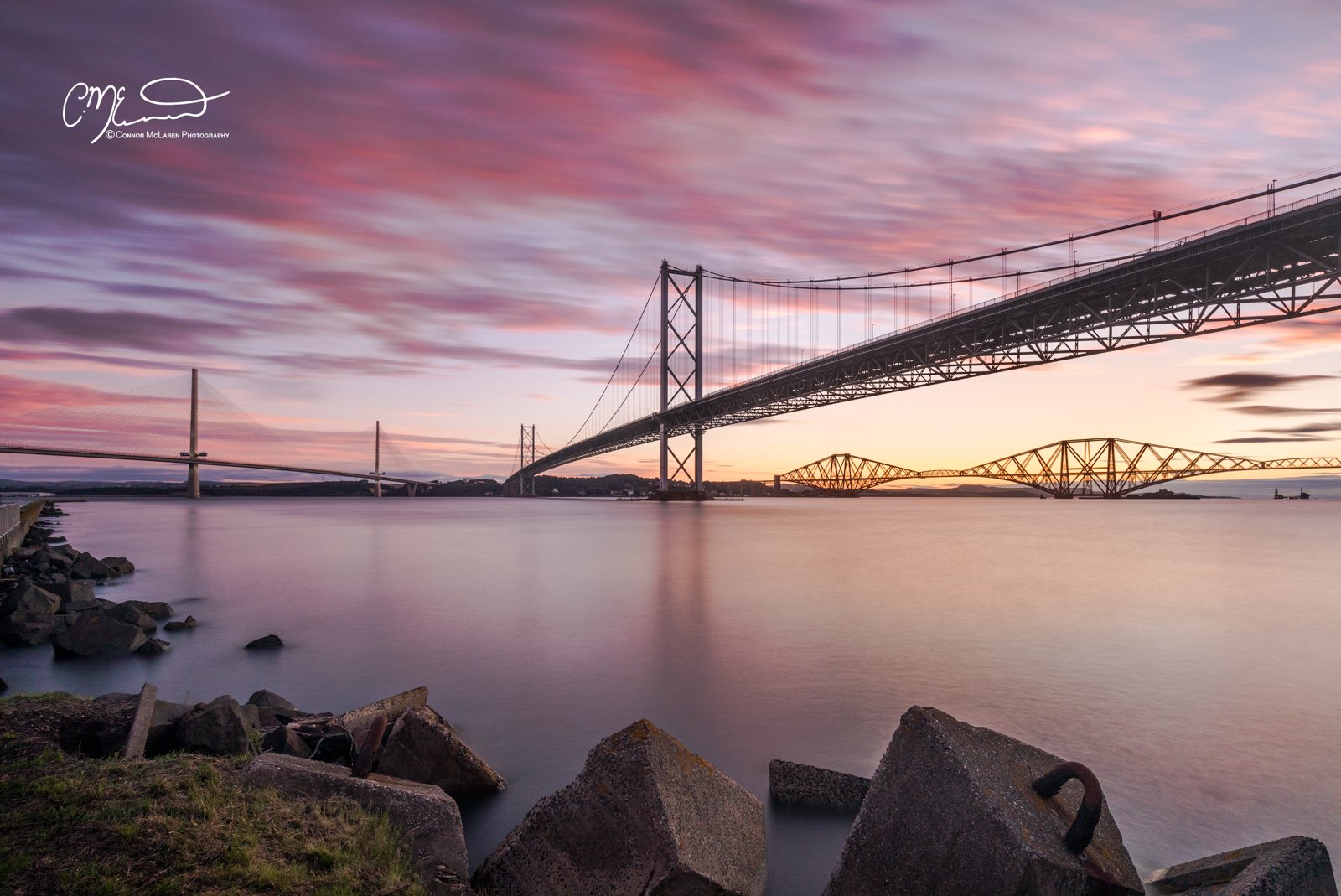 Scottish Lowlands, Scotland, Forth bridge Edinburgh. A long suspension bridge over a calm body of water during sunset, with a colorful sky featuring pink and purple clouds and rocks along the shoreline.