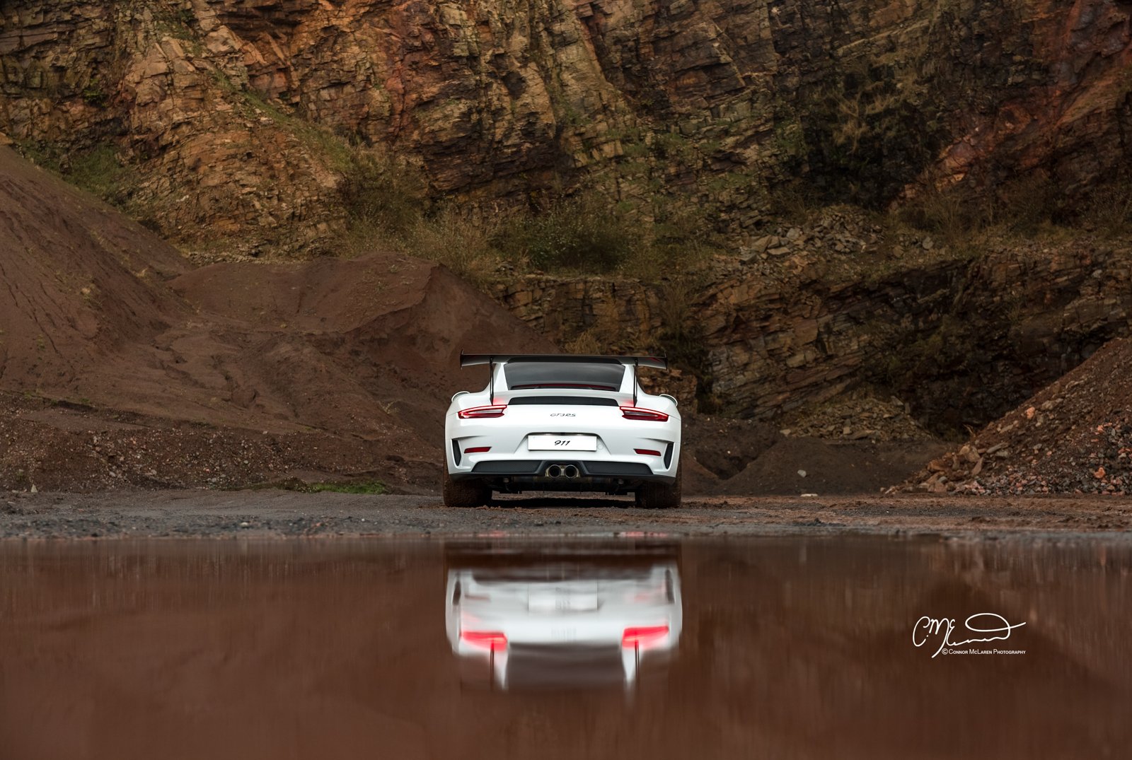 A white Porsche 911 GT3 RS parked in front of a rugged dirt landscape with rocky cliffs, with its reflection visible in a nearby shallow pool of water.