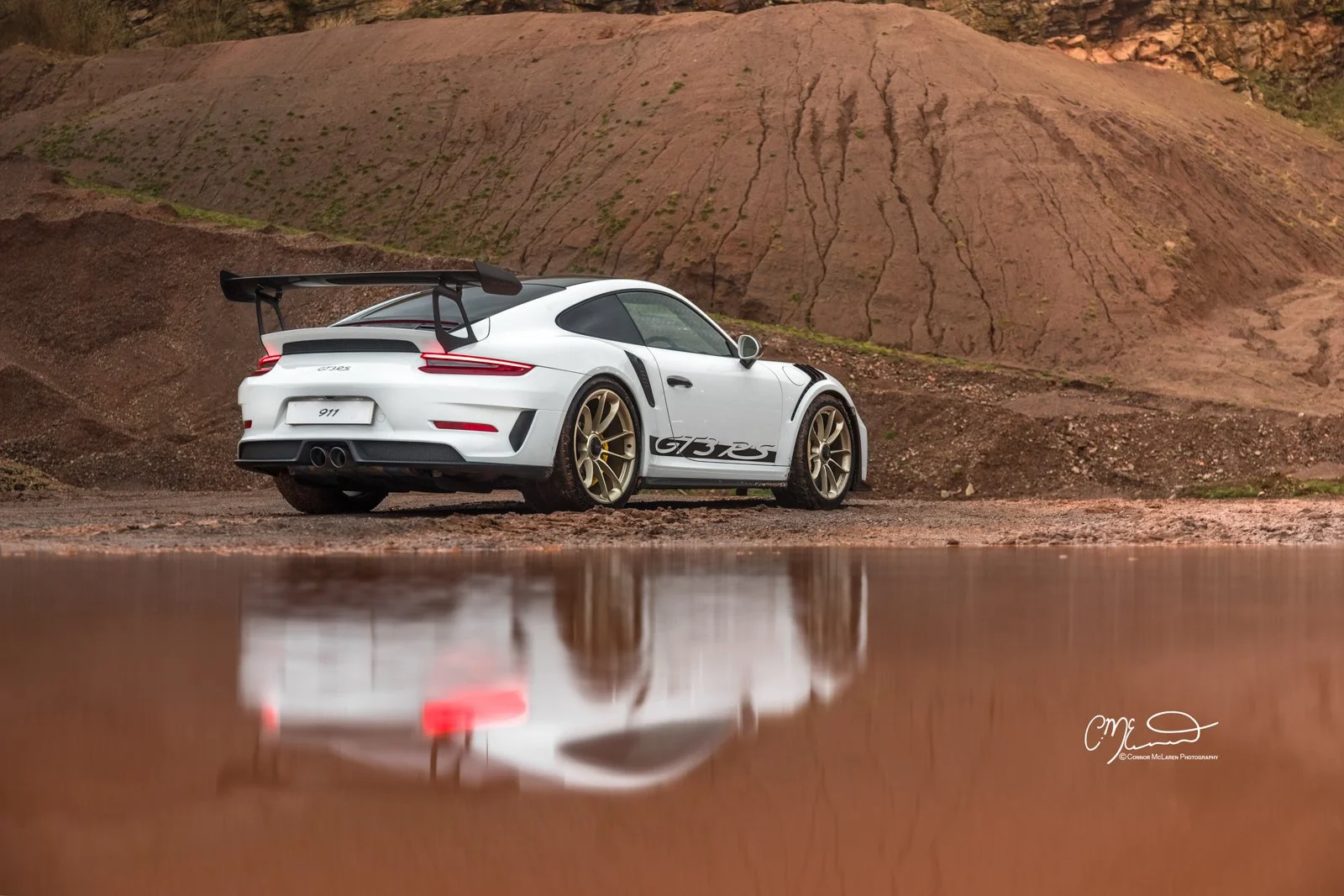 White Porsche 911 GT3 RS with gold rims reflecting in a pond against a rocky landscape.