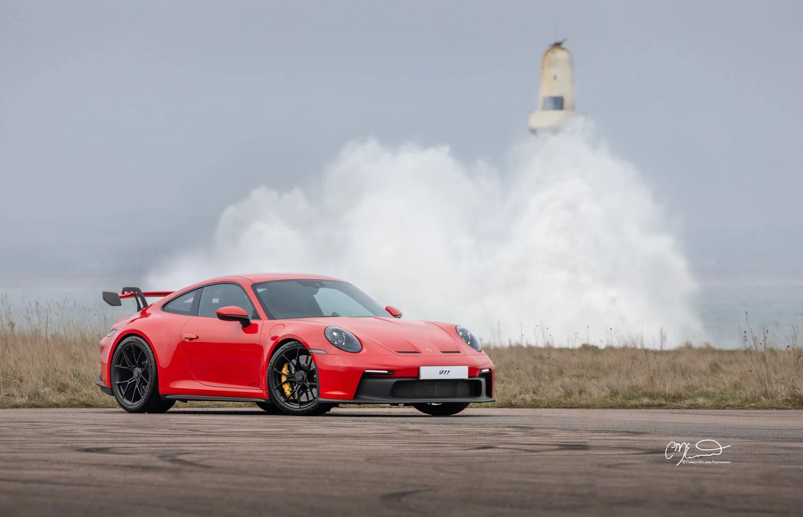 Red Porsche 911 GT3 parked on a tarmac with a lighthouse and ocean in the background, with a large splash of water behind the lighthouse.
