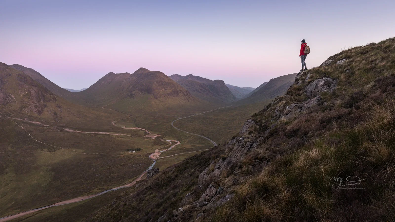 Glen Coe and Glen Etive, Western Highlands, Scotland. A person with a backpack standing on a rocky hillside overlooking a green valley with winding roads and mountains at sunset.