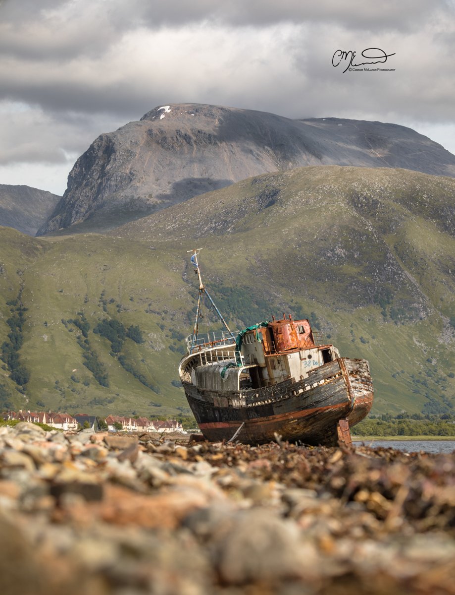 Corpach, Fort William, Ben Nevis, Western Highlands, Scotland. An abandoned, rusty boat resting on a rocky shore with a mountainous landscape in the background and partly cloudy skies.