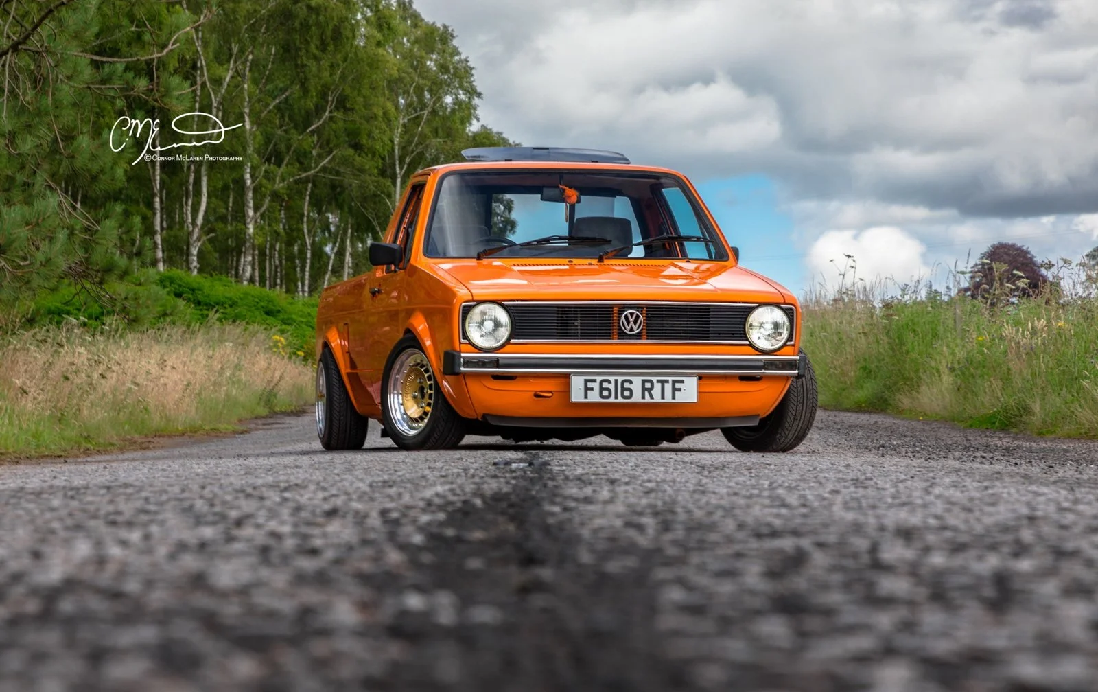Orange vintage Volkswagen Caddy pickup truck parked on an outdoor gravel road with lush green trees and cloudy sky in the background.