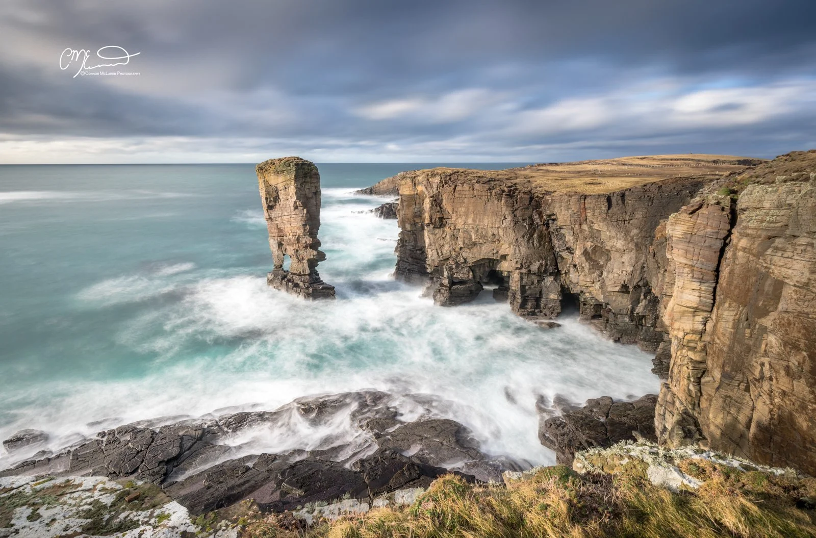Yesnaby, rainbow, Orkney, Scotland. Cliffs on a rugged coastline with a large sea stack in the water, stormy sky overhead, and waves crashing against rocks.