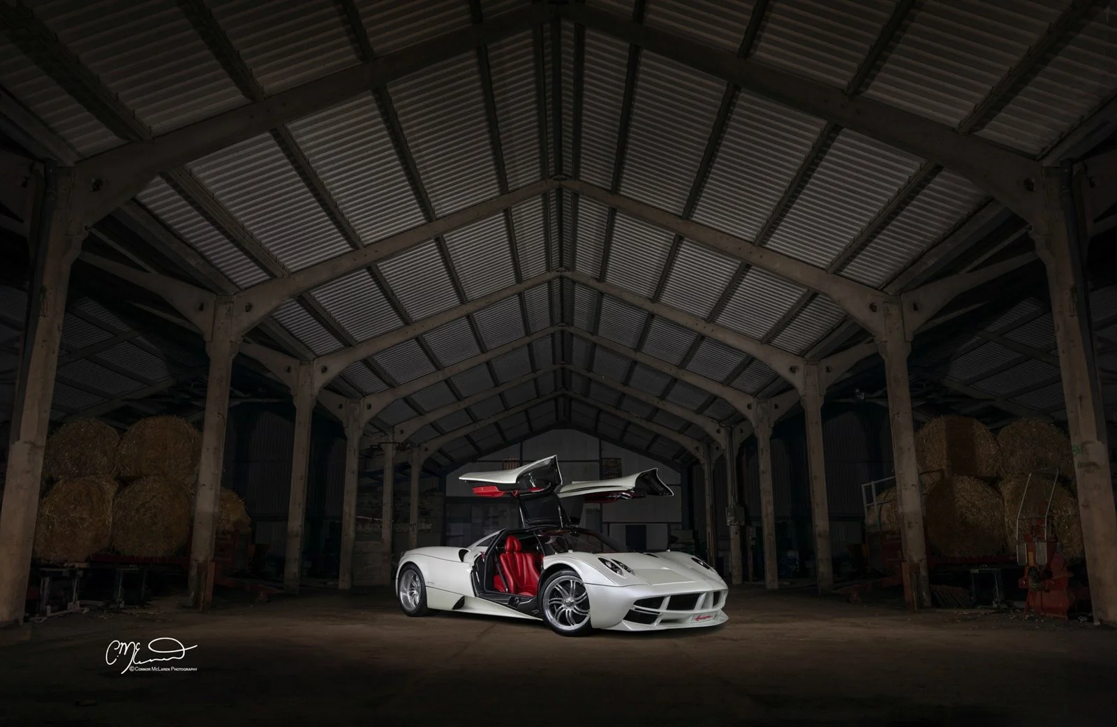 Pagani Huayra, white carbon fibre, red interior, barn. A white luxury sports car with a red interior in a barn with hay bales on the sides, with the car doors open vertically.