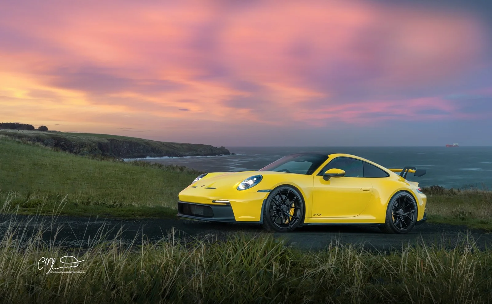 Yellow Porsche GT3 sports car parked on a scenic coastal road with grass and cliffs in the background, at sunset