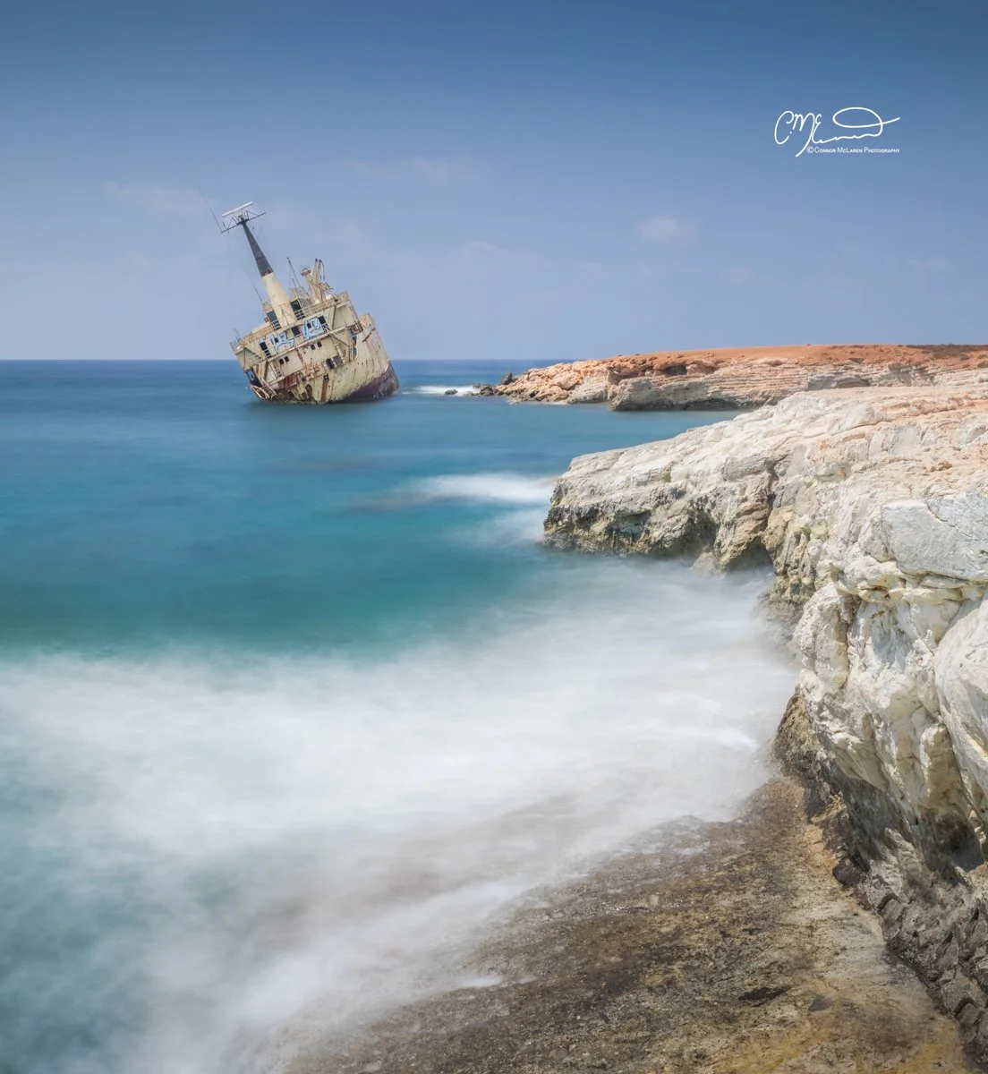 Edro shipwreck, Cyprus. A shipwreck partially submerged and leaning sideways in calm blue ocean waters near a rocky shore under a clear blue sky.