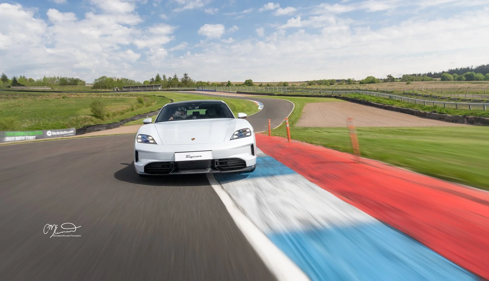A white Porsche Taycan sports car on a race track, approaching a turn, with colored curbs and a scenic landscape under a partly cloudy sky.
