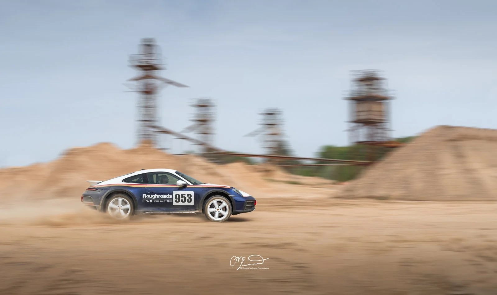 A racing Porsche 911 Dakar Roughroads with a blue, white, and red livery speeds through a dirt track, kicking up dust, with industrial structures in the background.
