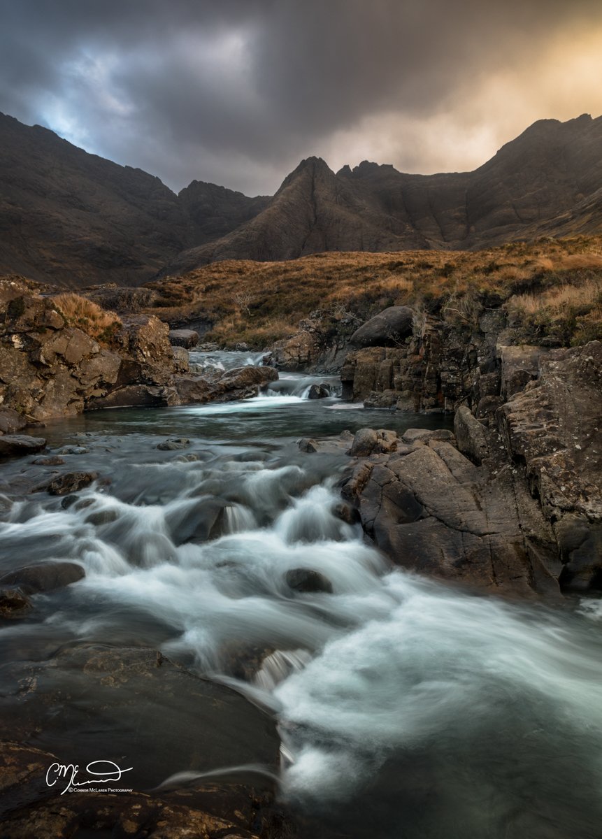 Scotland, Isle of Skye, Fairy Pools. A mountain stream flowing through rocky terrain with dark clouds overhead and rugged mountains in the background.