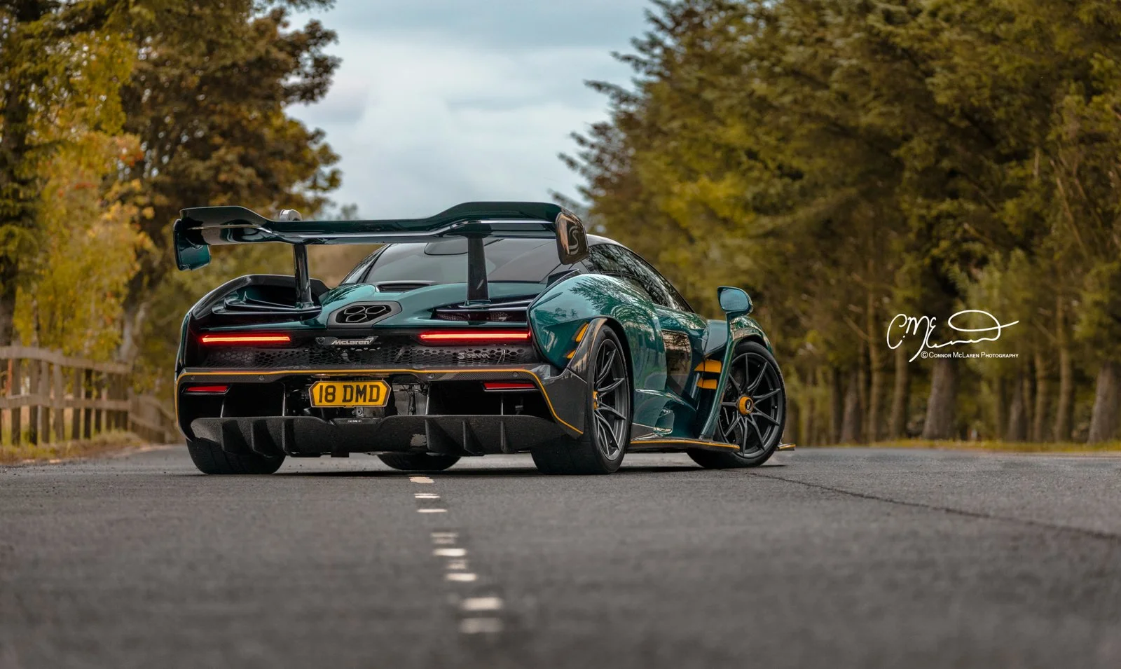 MSO McLaren Senna, full Carbon Green. A sleek black sports car with a large rear wing, racing tires, and yellow accents, parked on a rural road with trees on both sides.