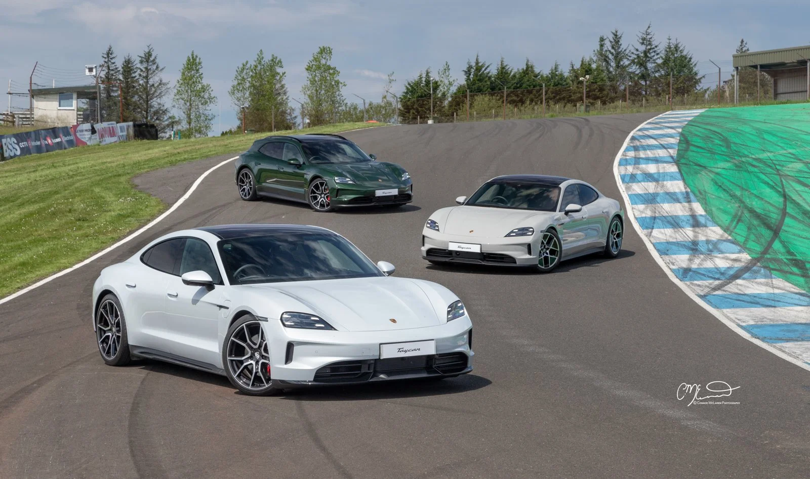 Four Porsche cars, white, silver, dark green, and white, parked on a racetrack with a curving blue and white striped section and green runoff area, surrounded by trees and structures.