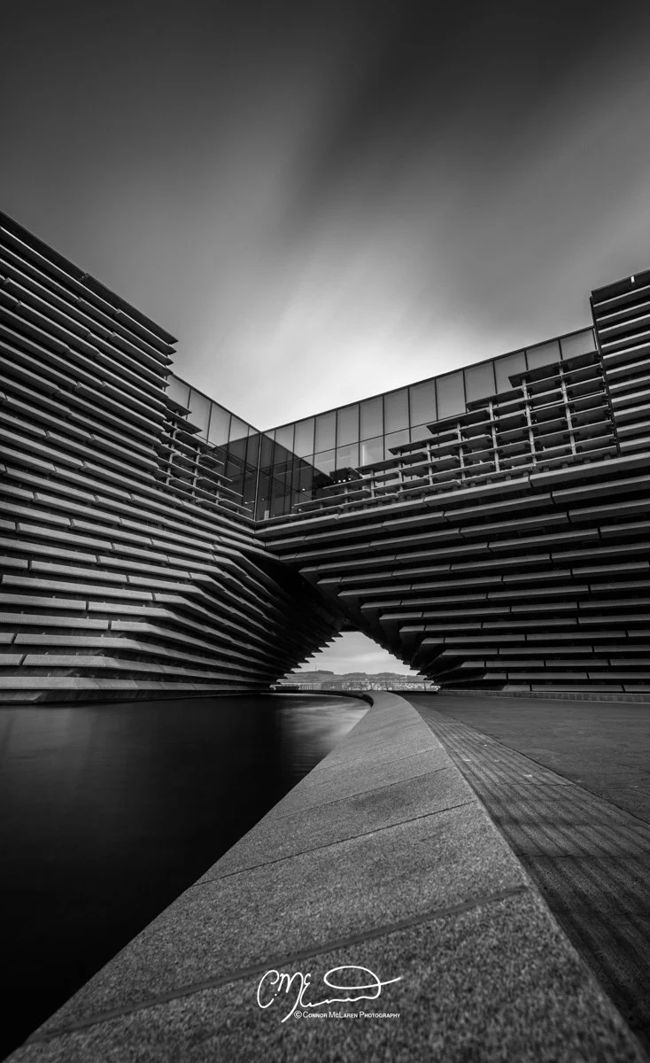 Scotland, Dundee, V&A. Black and white photo of modern architectural building with angular design, glass windows, and layered exterior walls reflected in a water feature.