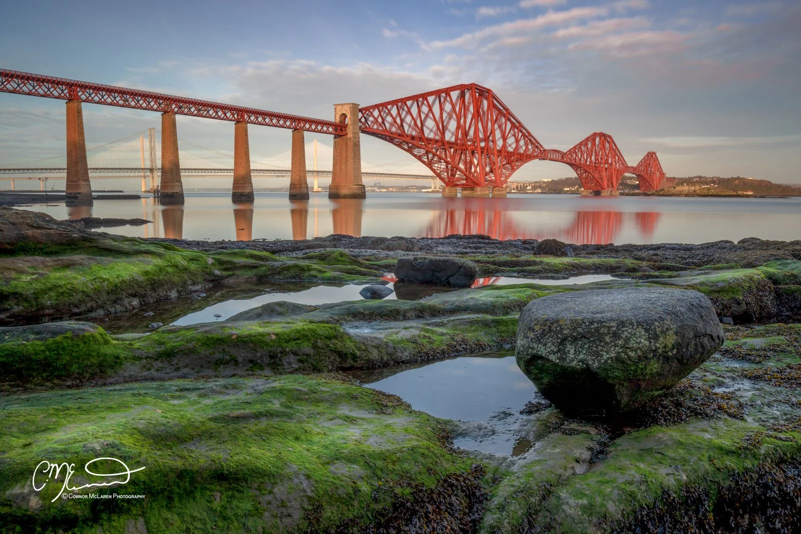 Scottish Lowlands, Scotland, Forth bridge Edinburgh. Red bridge over calm water with rocky, moss-covered shores in foreground and a partly cloudy sky at sunset
