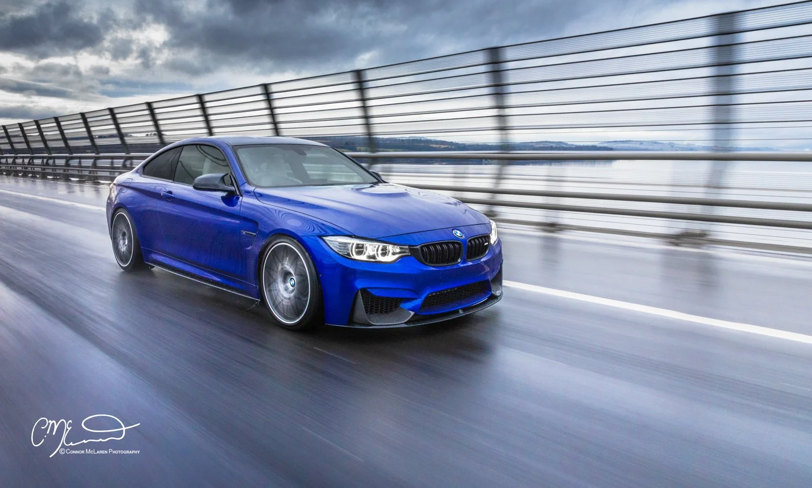 A blue BMW M4 sports car driving on a wet roadway with a metal barrier on the side, under a cloudy sky.