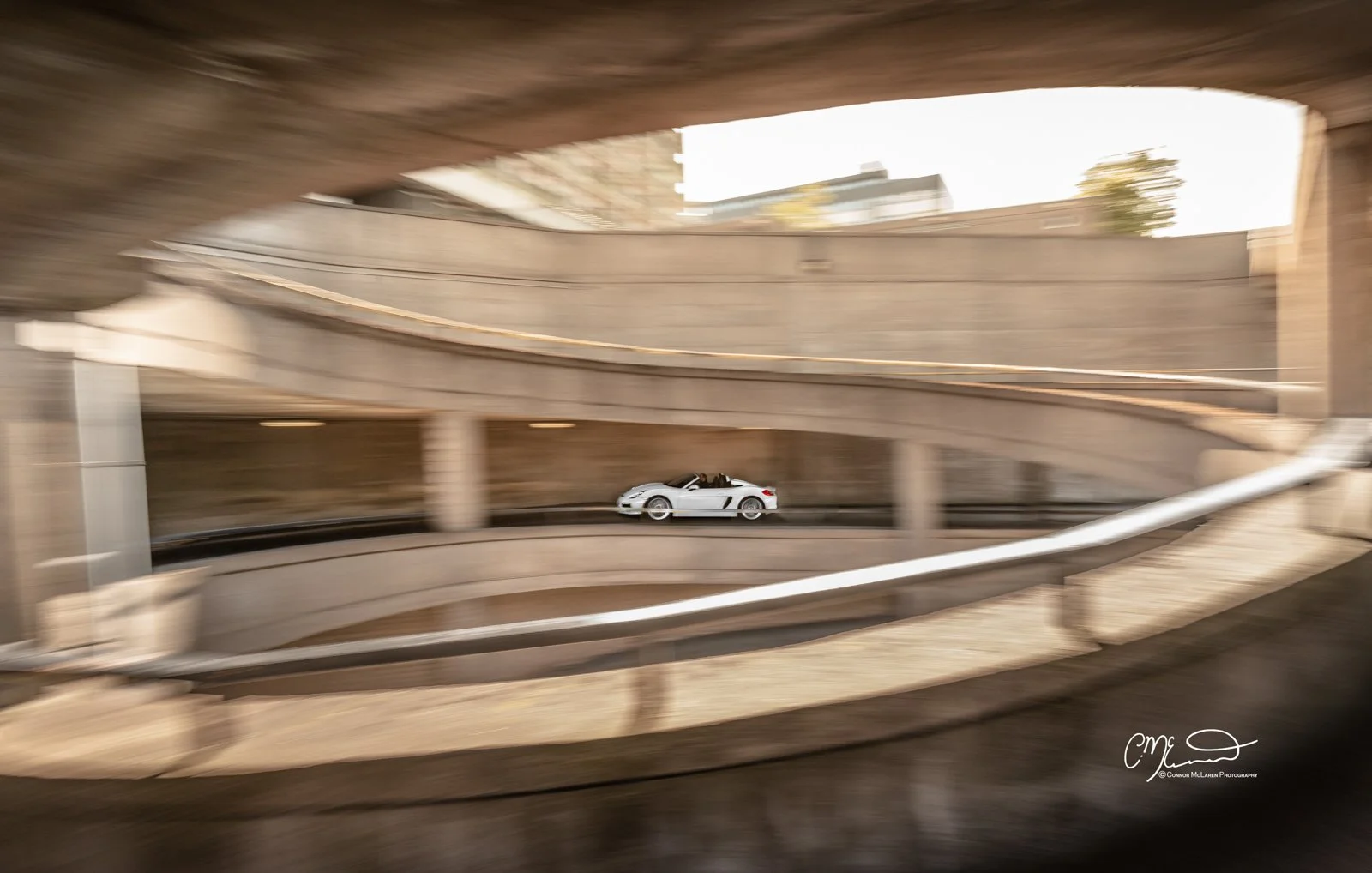 A white Porsche Boxster Spyder sports car driving on a spiral ramp within a concrete parking garage.