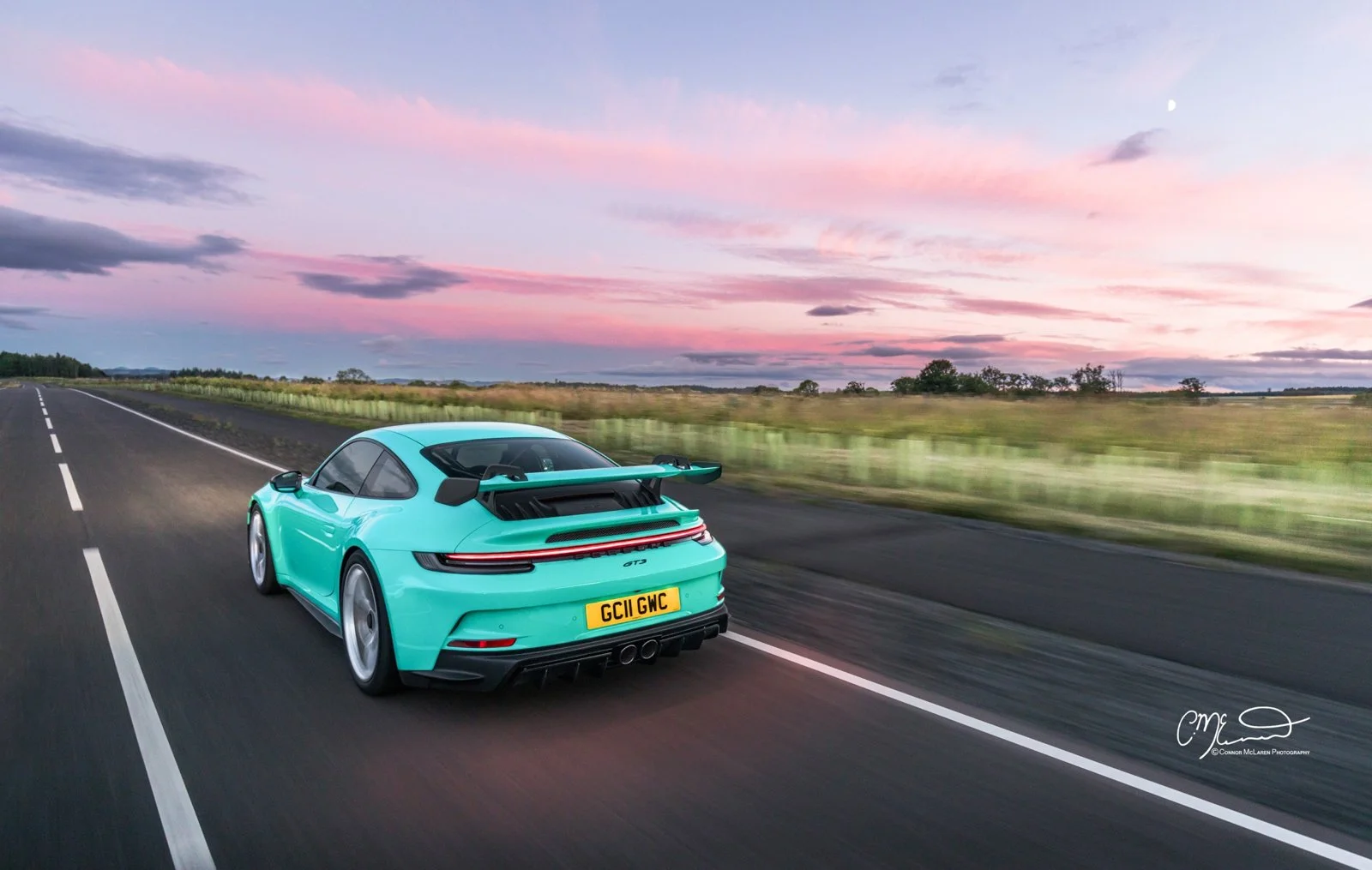 Mint Porsche 911 GT3 992. A turquoise Porsche 911 sports car driving on an open road during sunset with pink and purple clouds and a visible moon in the sky.