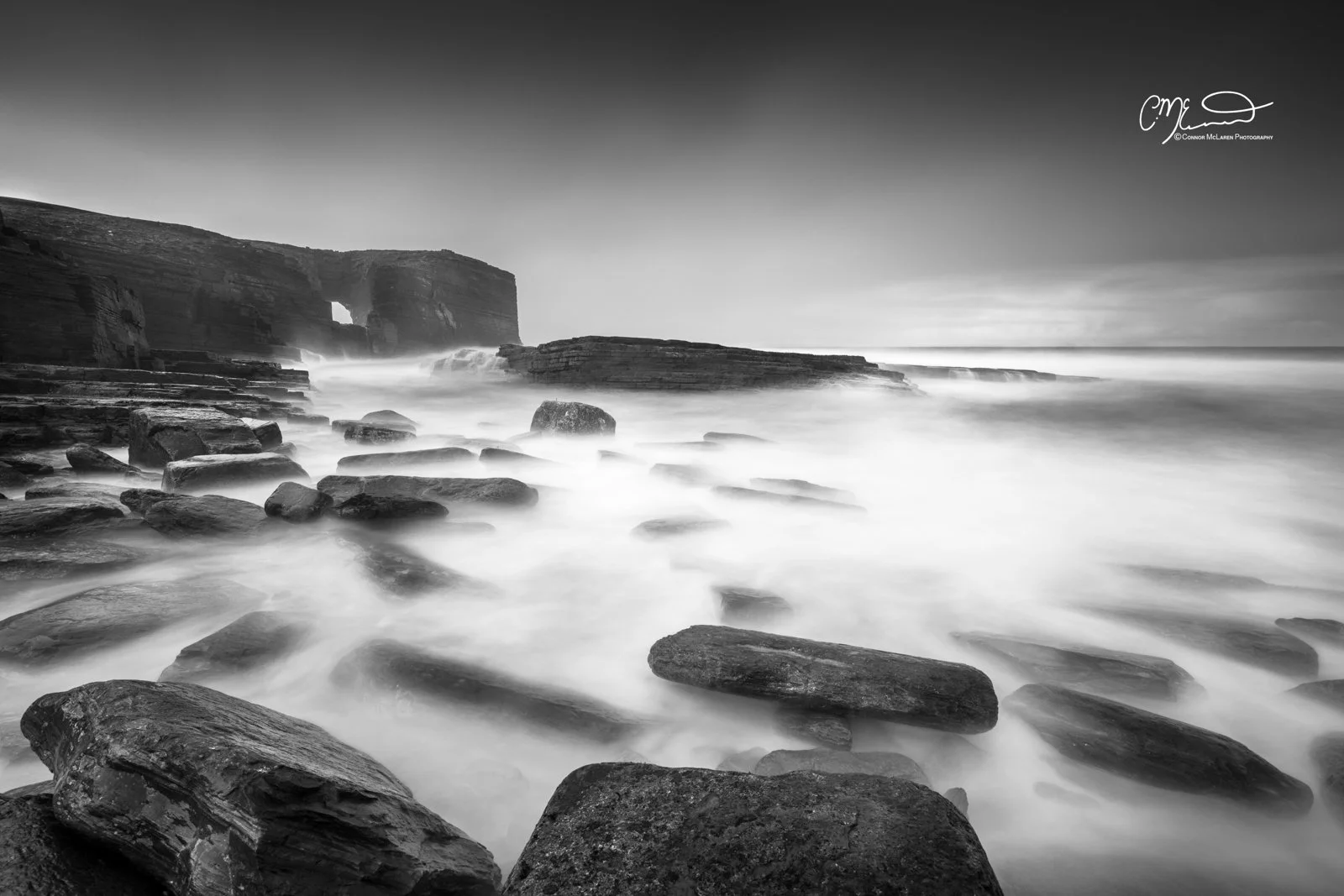 Scotland, Orkney. Black and white long exposure photograph of rocky coastline with cliffs in the distance and ocean waves washing over rocks.