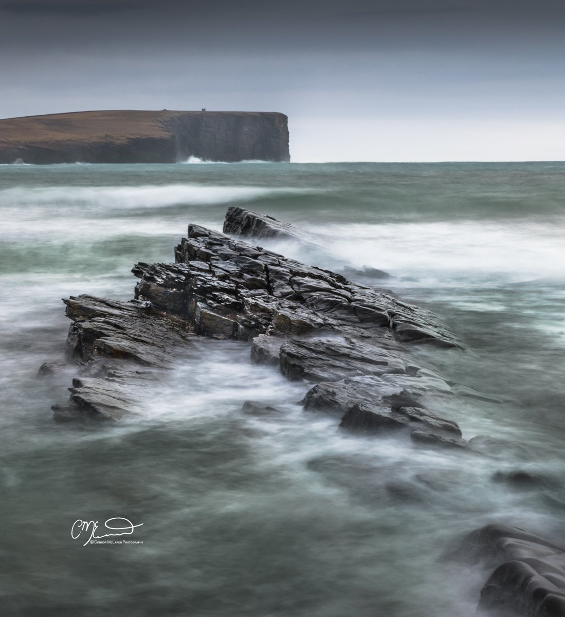 Orkney, Scotland. Rock formations in the ocean with waves crashing, and cliffs in the background under a dark, cloudy sky.