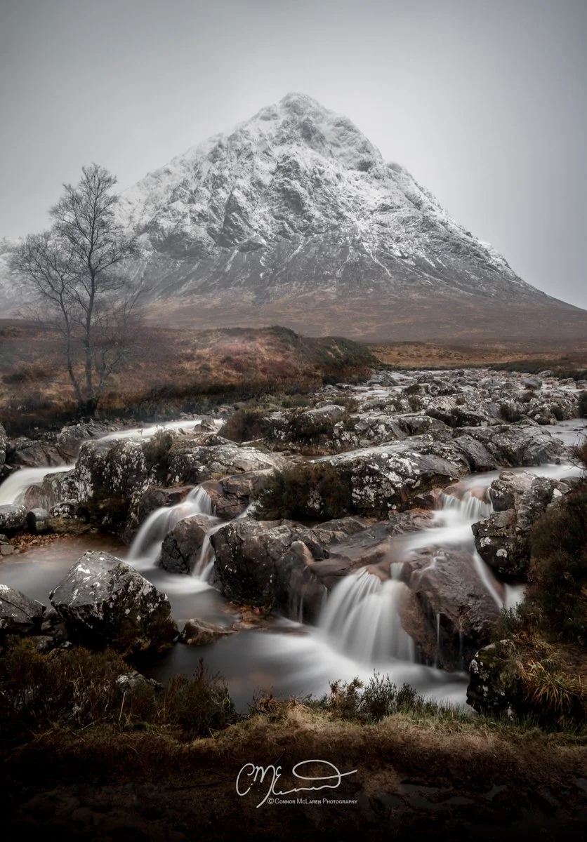 Glen Coe and Glen Etive, Western Highlands, Scotland. Snow-capped mountain with a rocky stream in the foreground and leafless trees to the left.