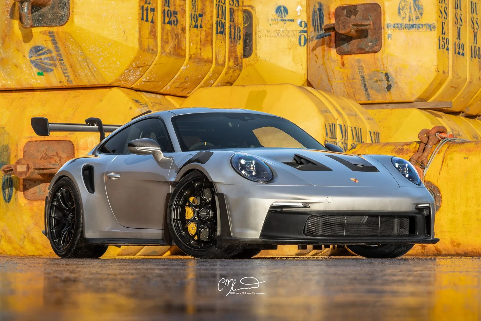 Silver Porsche 911 GT3 RS race car parked in front of large yellow industrial containers with rust and markings.