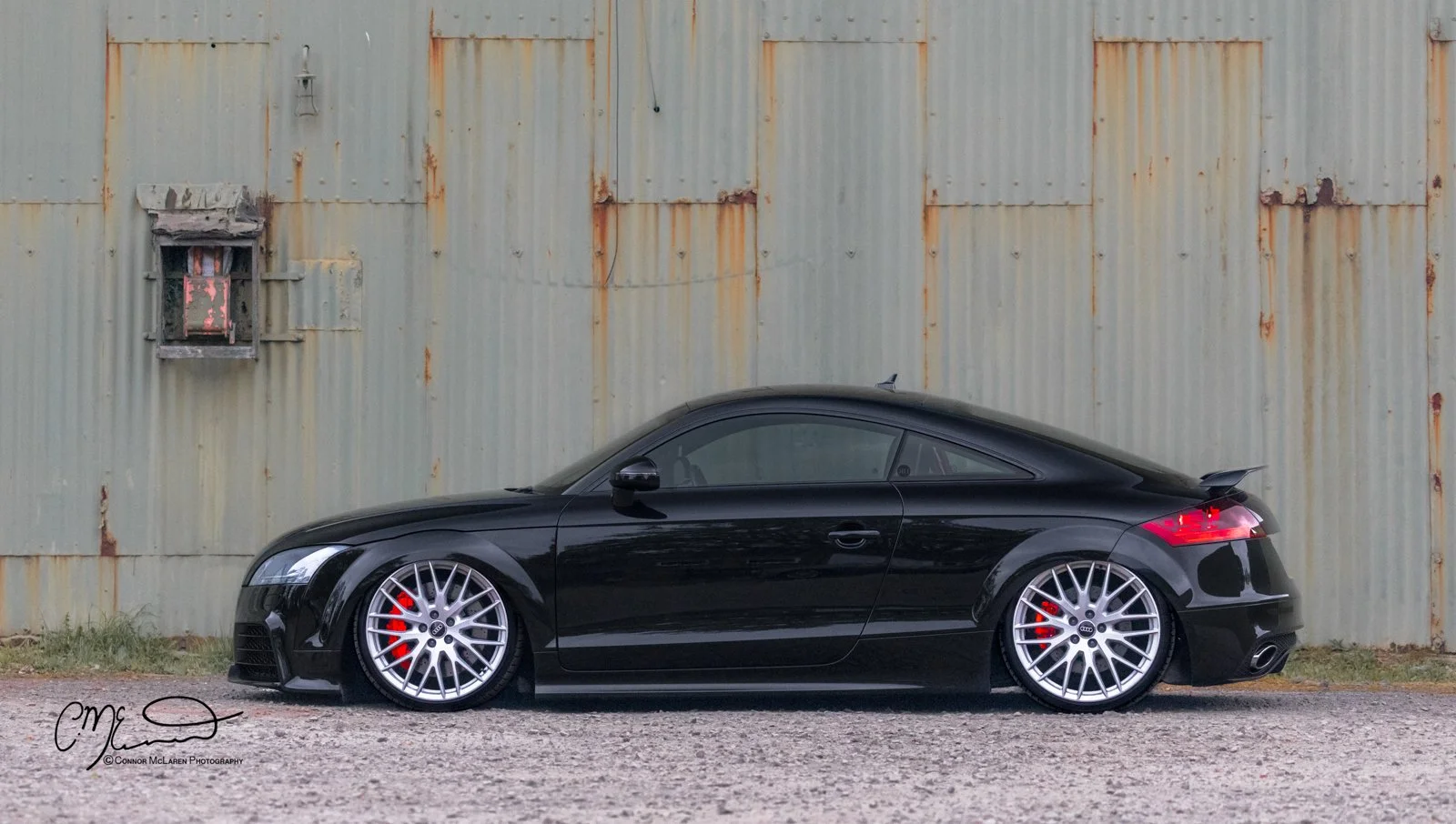 Custom Audi TTRS in black, bagged air suspension. A black Audi TT coupe parked on gravel in front of a weathered, rusted metal wall with a small boarded-up window and an emergency service box.