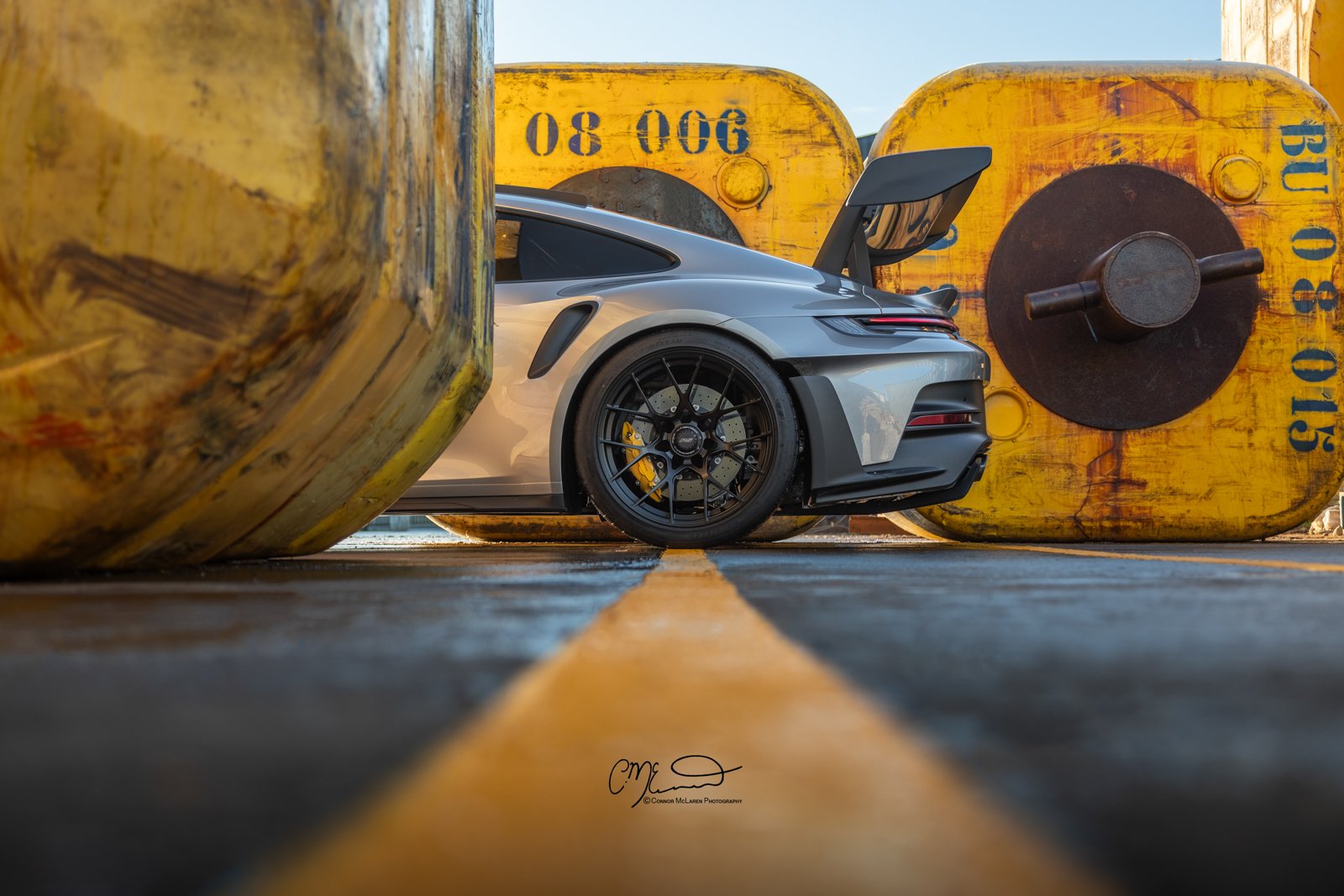 A silver Porsche 911 GT3 RS sports car with a large rear wing parked between large yellow industrial spools in an outdoor setting.