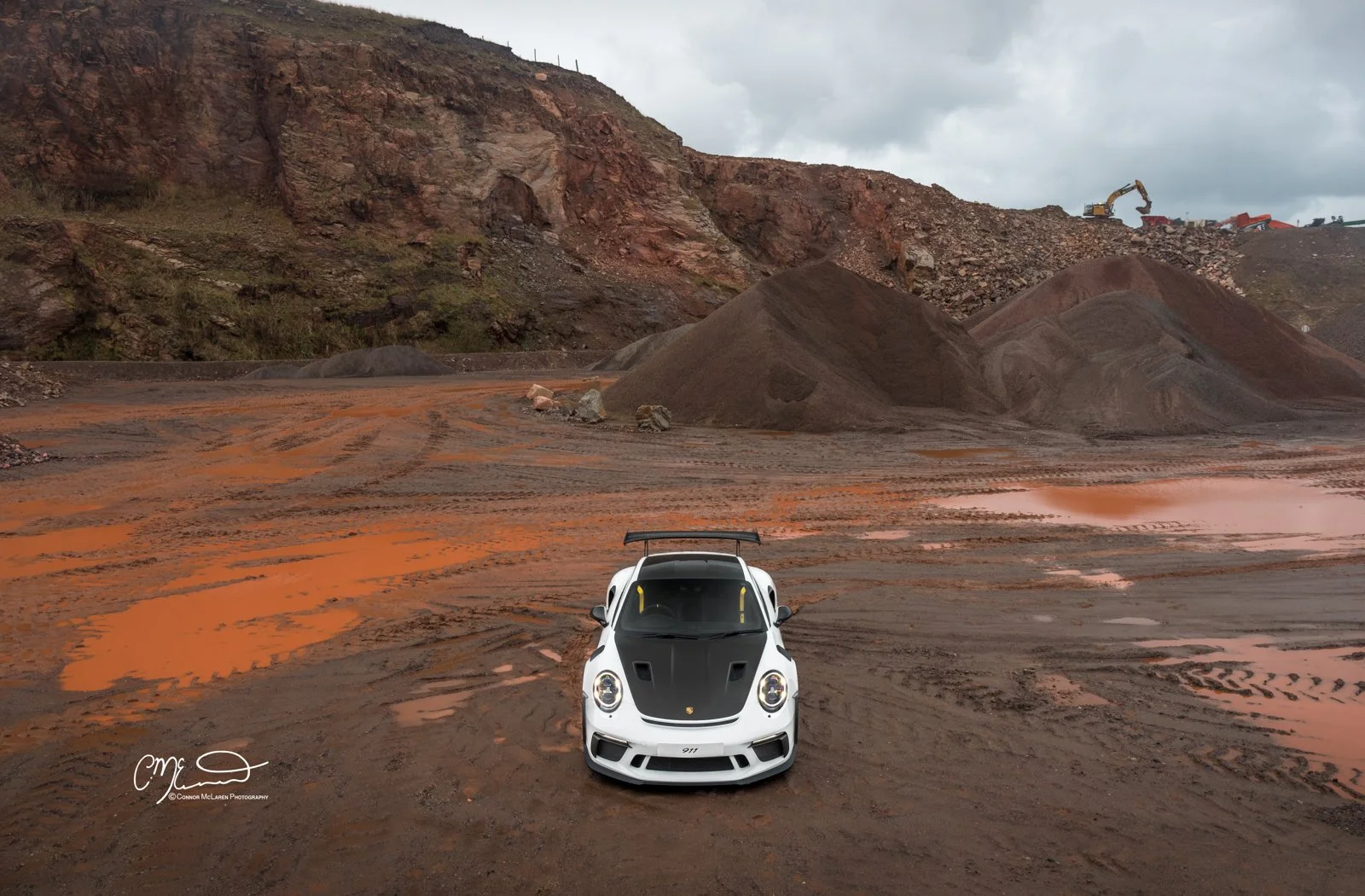 A white Porsche 911 GT3 RS sports car with a black hood and rear wing parked on a muddy construction site or quarry with mounds of excavated dirt and a large excavator in the background under a cloudy sky.