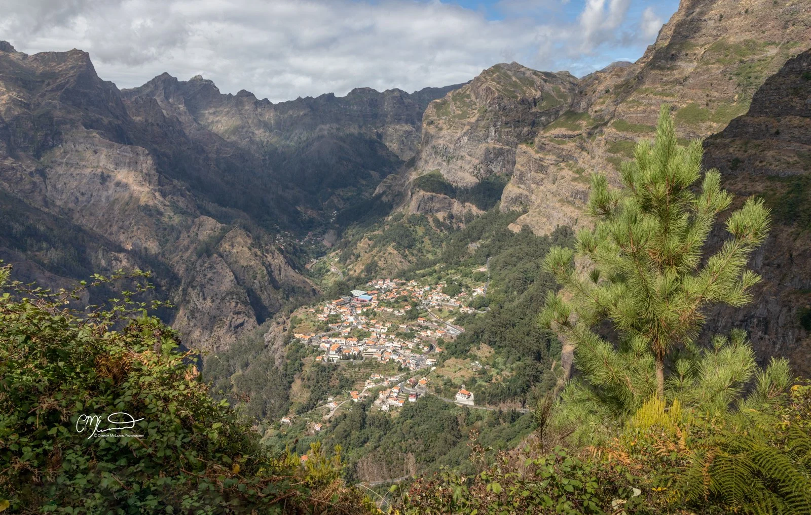 the nuns valley, curral das freiras, Madeira Portugal. A view of a small town nestled in a valley surrounded by steep, rugged mountains with sparse vegetation, some greenery, and a cloudy sky.