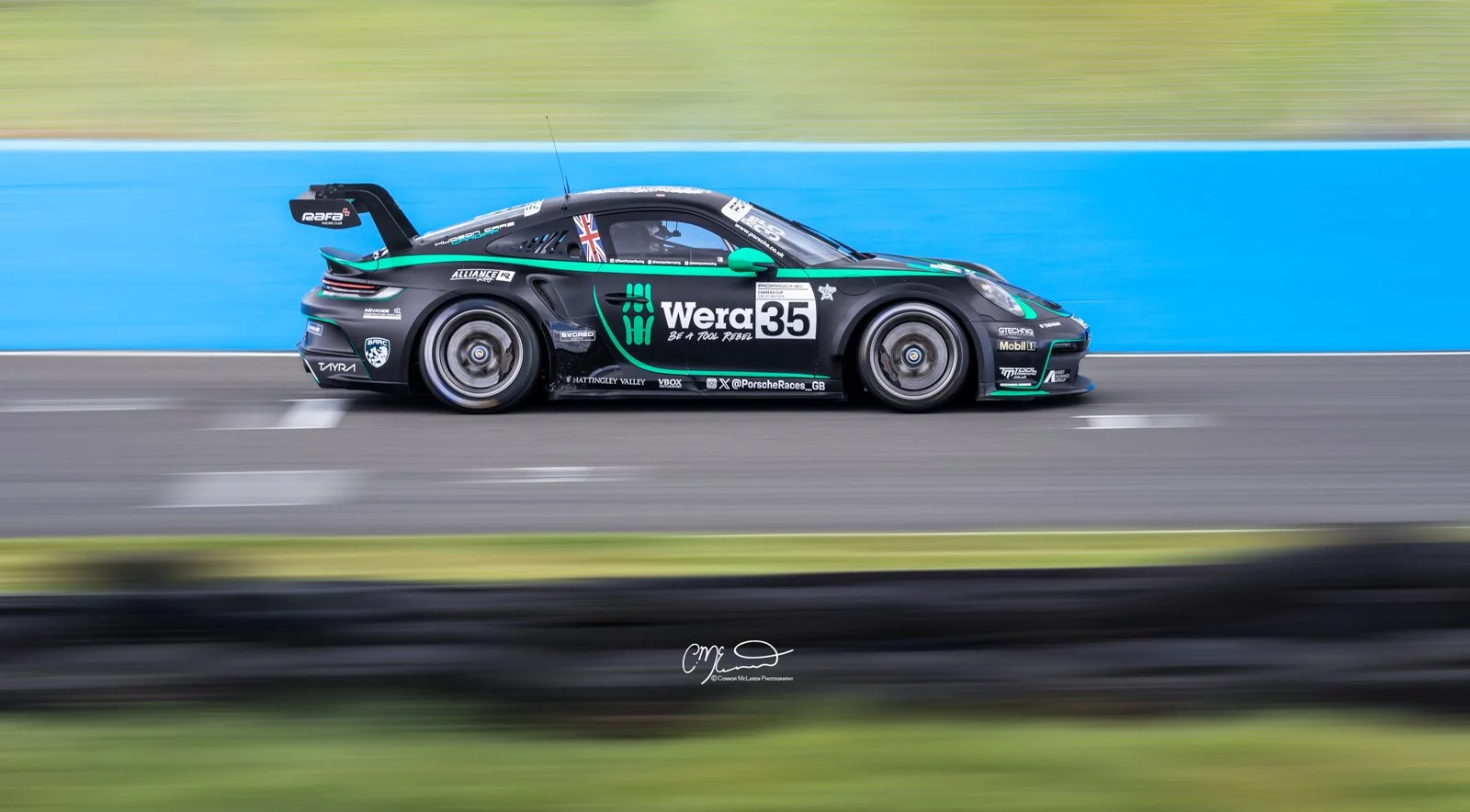 Porsche 911 GT3 Cup car, racing in the Carrera Cup at Knockhill racing. A black race car with various sponsor logos, including Wera, racing on a track with a blue wall in the background. The car has a rear wing and appears to be moving at high speed.