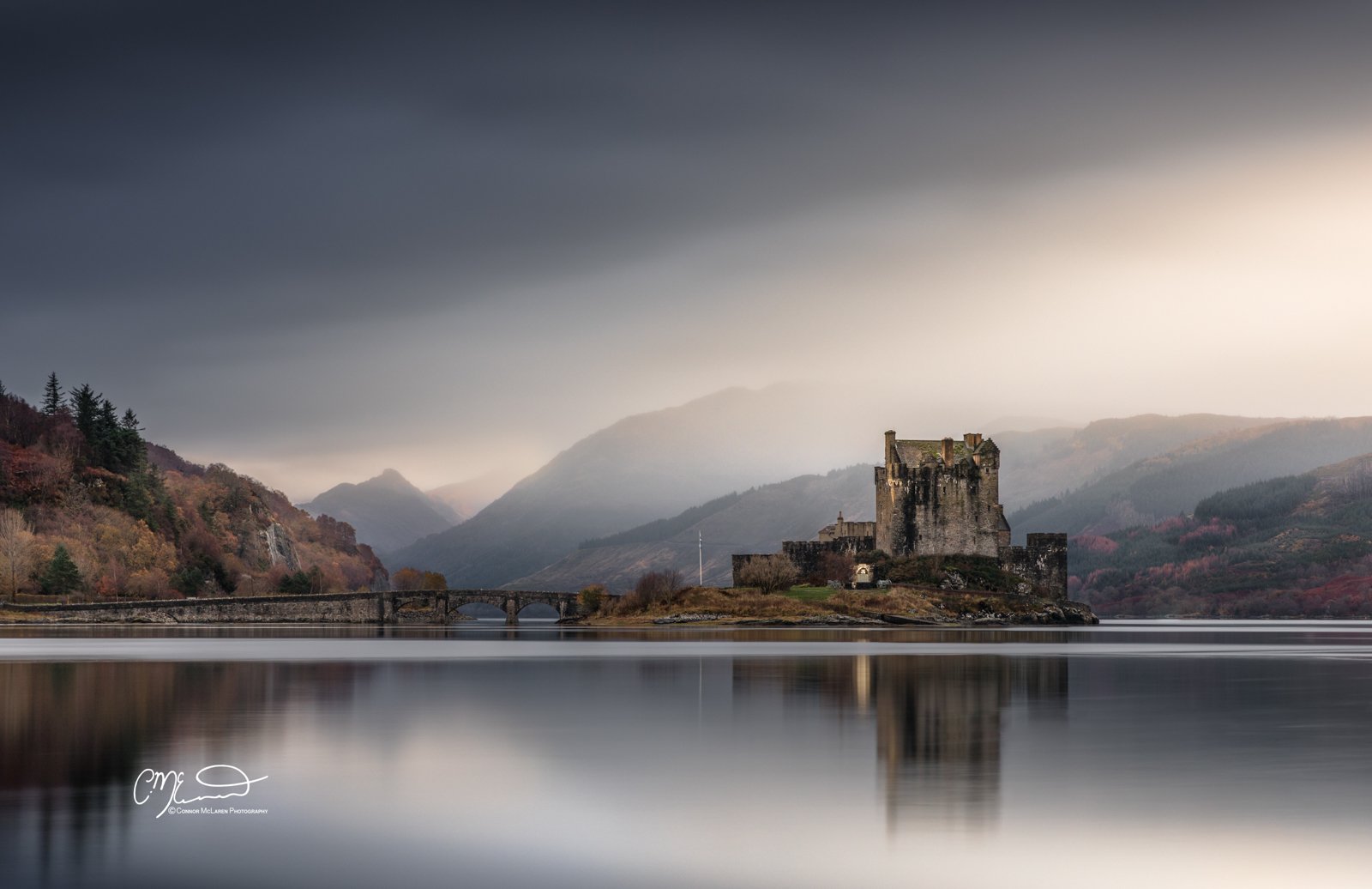 Bucket list locations, Scotland, Eilean Donan Castle. A historic castle on a small island surrounded by calm lake waters, with a background of misty mountains and a partly cloudy sky.