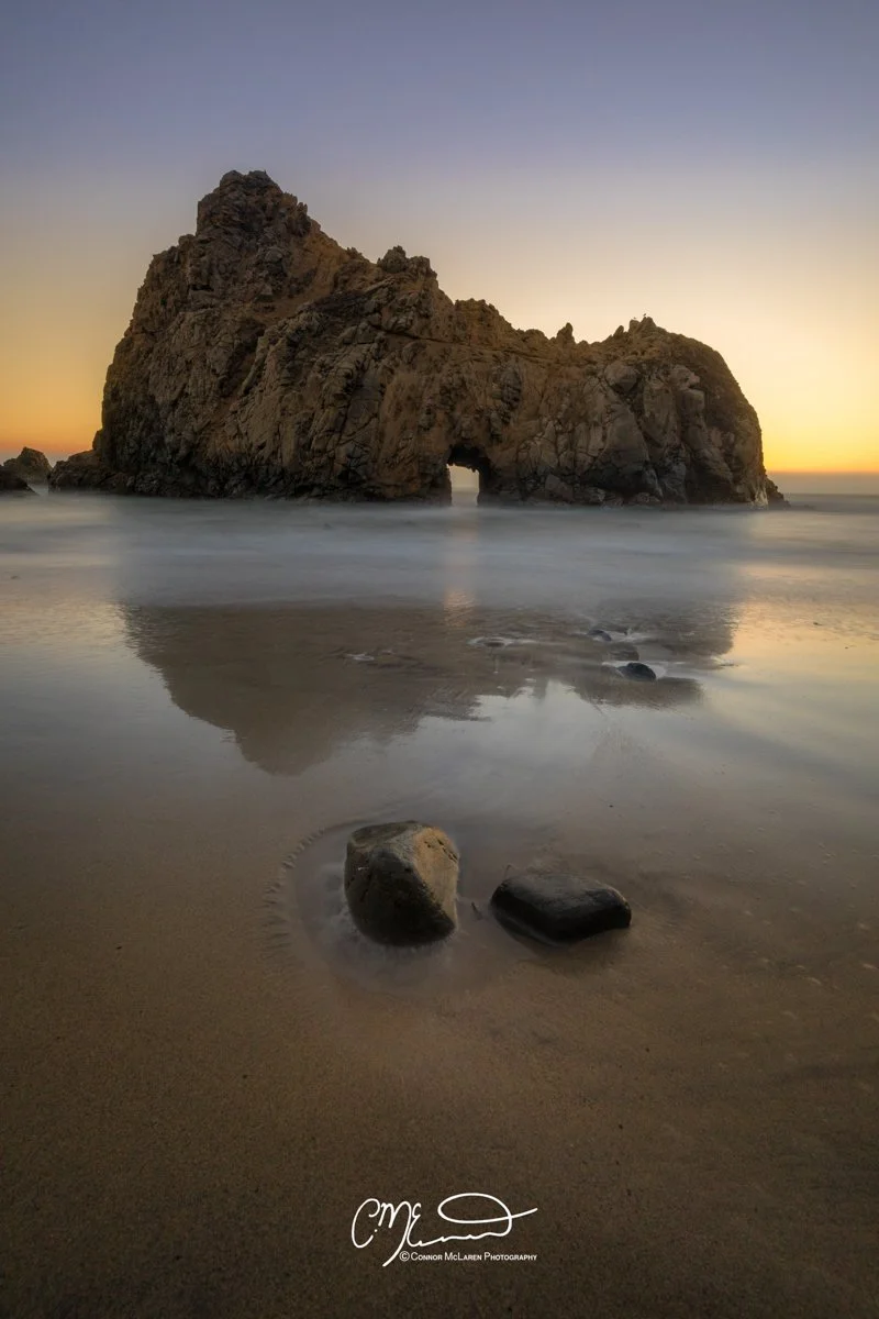 big sur, california, united states of america, usa. A large sea cave rock formation on a beach at sunset, with rocks in the foreground and their reflection in the wet sand.