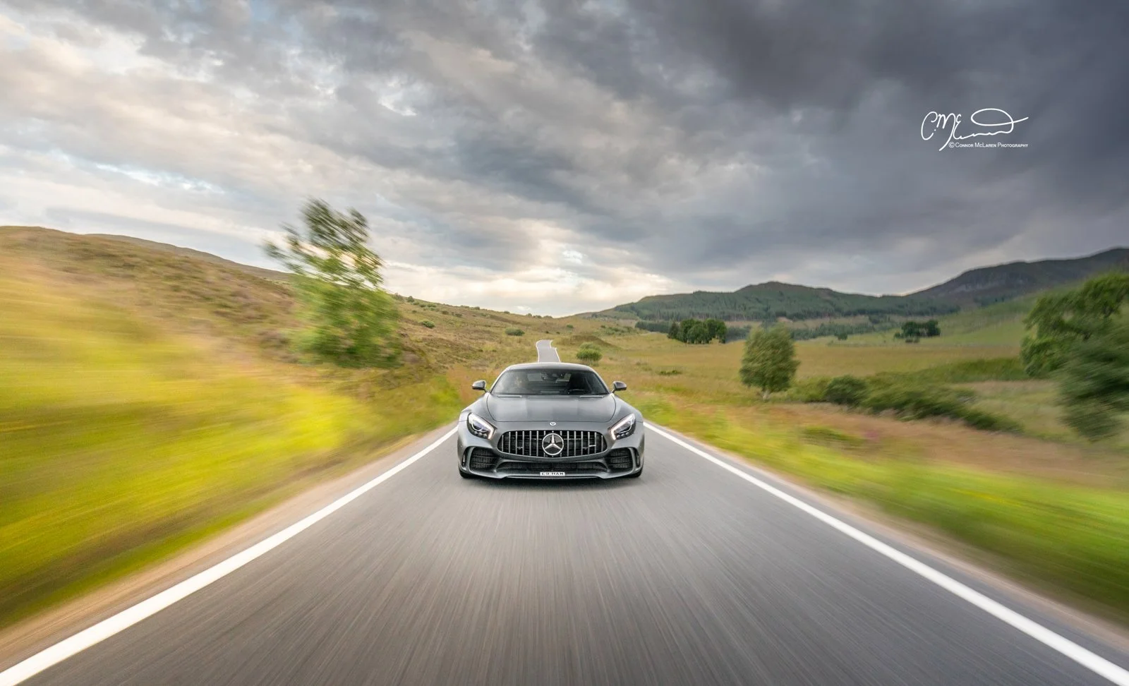 A sleek gray Mercedes-Benz AMG GT sports car driving down a two-lane rural road through green hills and mountains with a cloudy sky overhead.
