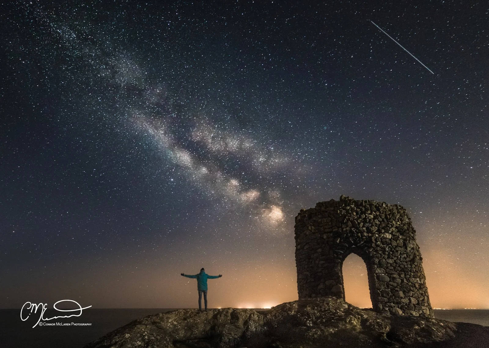 fife, Night Sky, Scotland.  Person standing with arms outstretched on a rocky landscape near a stone arch, looking at the starry night sky with the Milky Way galaxy and a bright meteor streaking through.