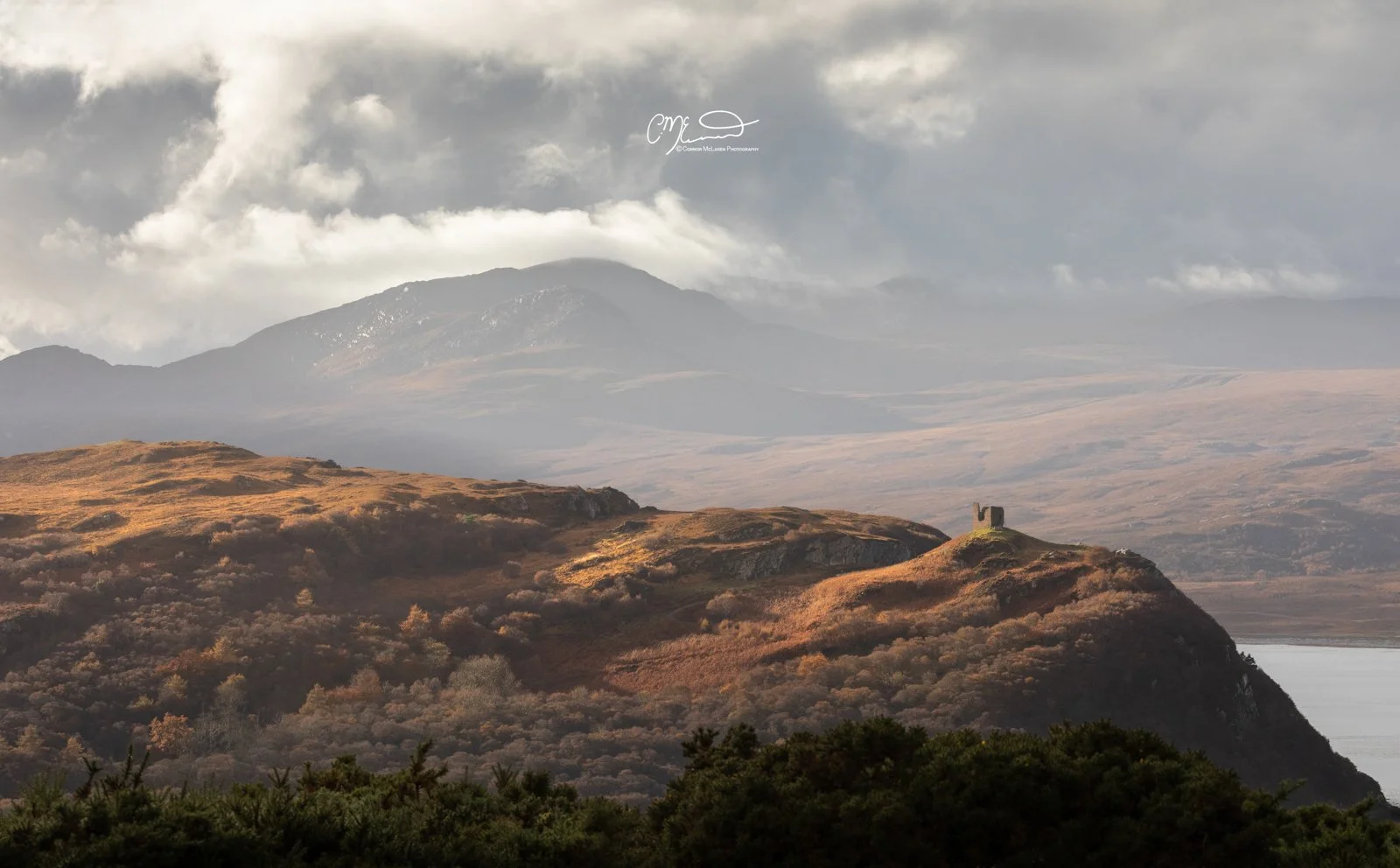 Scotland, north coast 500, NC500, castle varrich. Scenic landscape of hills and mountains with a small castle ruin on a hill, lush green trees in the foreground, and cloudy sky overhead.