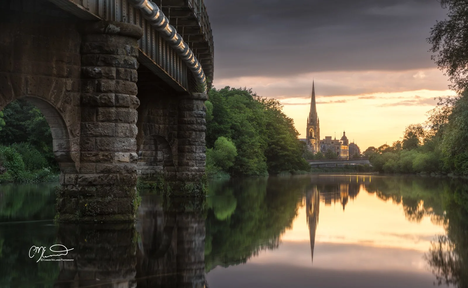 Perth Scotland, Scottish central highlands. View of a river at sunset with a stone bridge on the left, lush green trees on both sides, and a church with a tall spire in the background, reflected in the calm water.