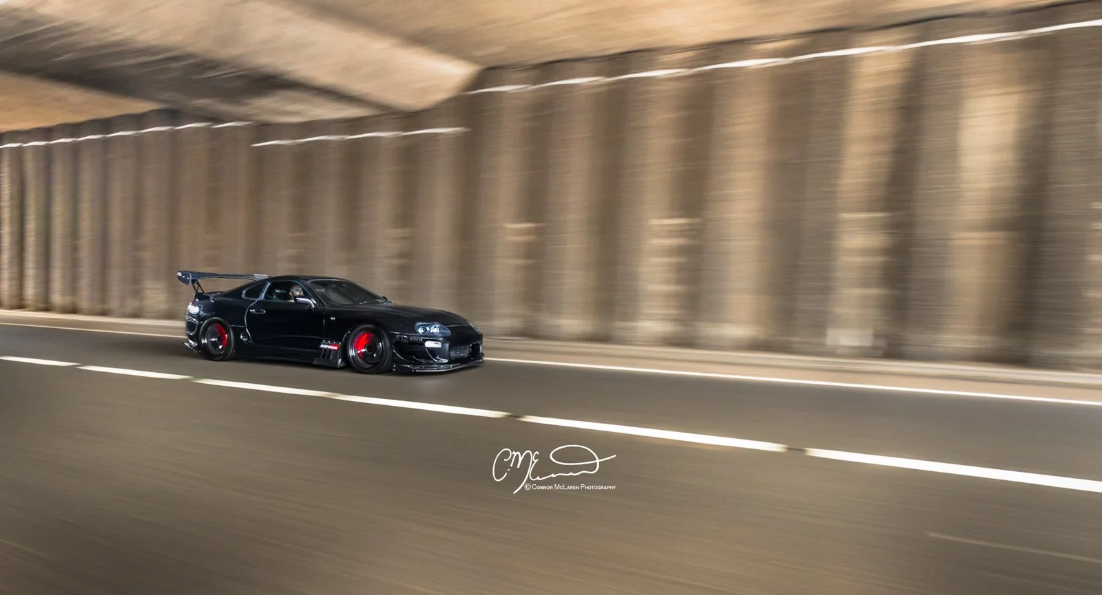 Custom Modified Toyota Supra 2JZ. A black sports car with a large rear wing and red accents on the wheels speeds through a tunnel, with blurred background indicating motion.