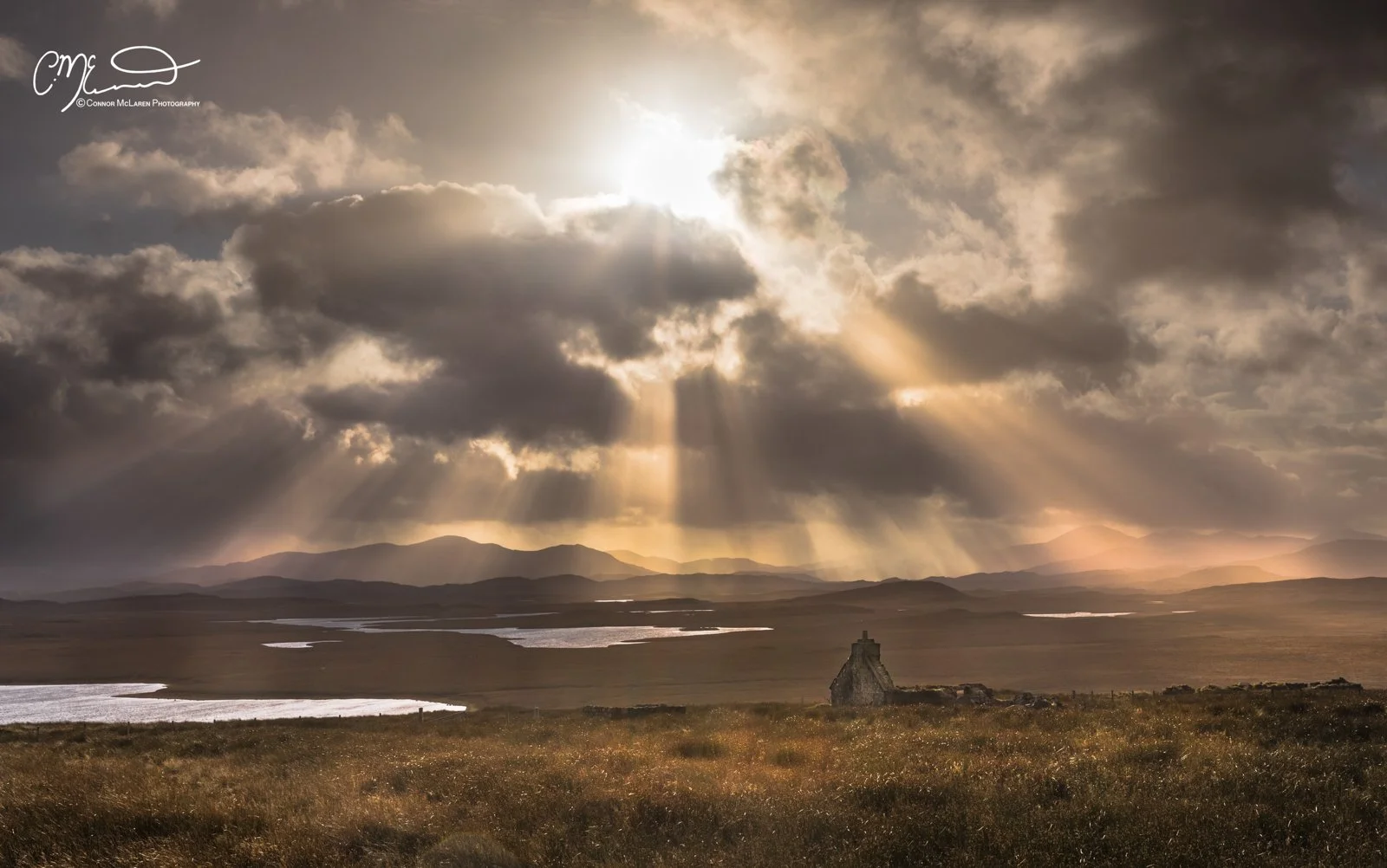 Scotland, Lewis Harris & Outer Hebrides, Stornoway. A landscape scene of a field with a small stone structure, bodies of water, and distant mountains under dramatic cloudy skies with sunlight rays breaking through.