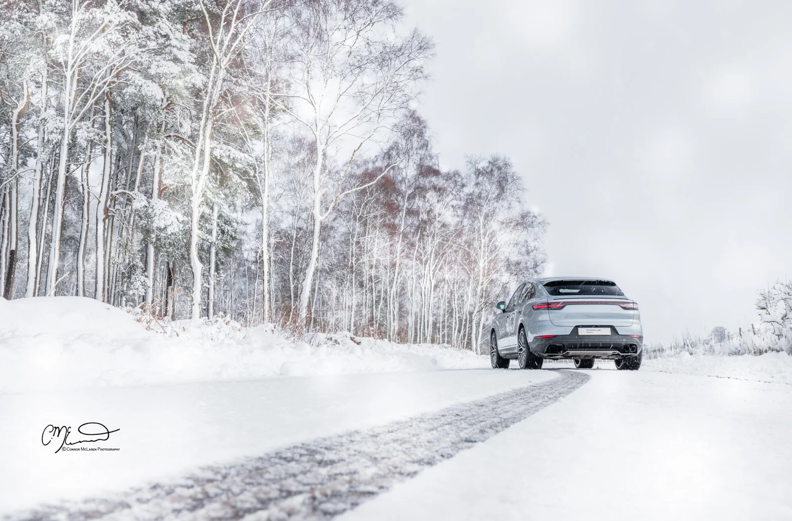 A silver Porsche Cayenne coupe car parked on a snow-covered road next to a snowy forest with leafless trees.