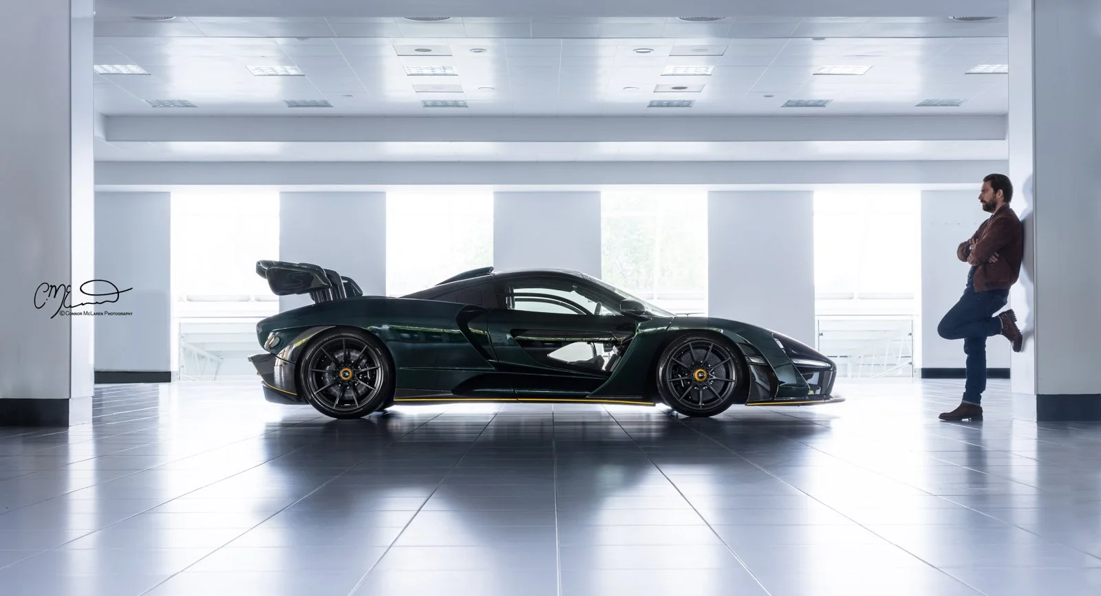 Mr JWW McLaren Senna MSO full carbon. A dark green hypercar with black wheels is parked indoors, with a man leaning against the wall nearby, in a modern, brightly lit room with tiled floors and large windows.